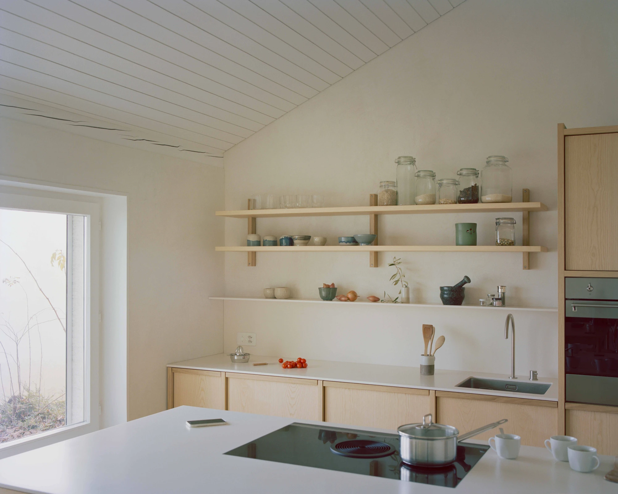 Family Villa by Noue Studio — The kitchen workspace centers on a green-glazed induction cooktop set into a white composite countertop, flanked by white ceramic vessels and stainless...