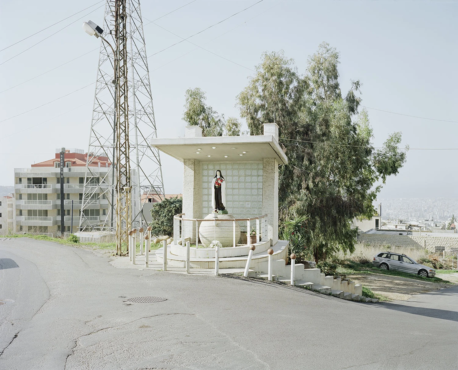 ASNAM (Idol Statues) by Georges Salameh — A white plaster statue of the Virgin Mary stands in a small shrine structure with rendered walls and a flat canopy, elevated on a concrete pedestal within a...