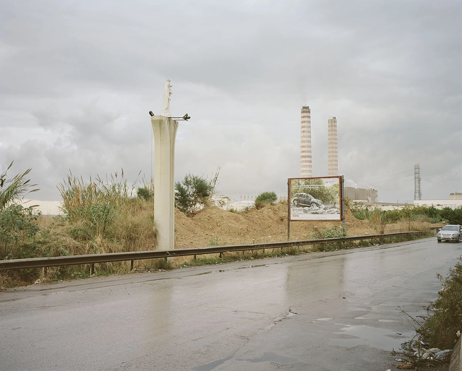 ASNAM (Idol Statues) by Georges Salameh — A monumental white marble or plaster statue in classical Marian pose—crown-topped, arms extended—rises from a weathered stone pedestal along a wet asphalt road.