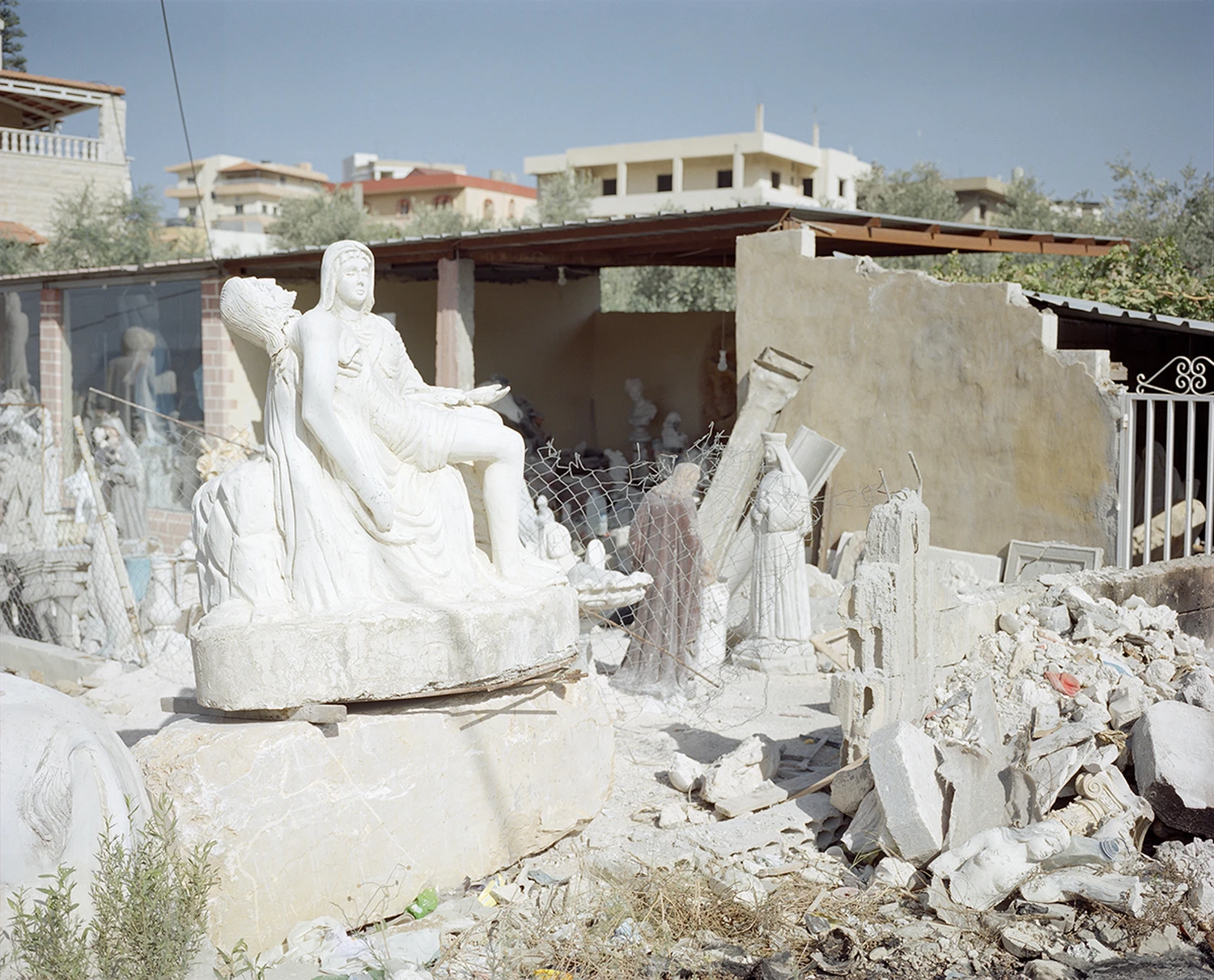 ASNAM (Idol Statues) by Georges Salameh — A monolithic marble statue of the Virgin Mary dominates a stone-working yard in Lebanon, her seated figure rendered in classical pose with crown and...