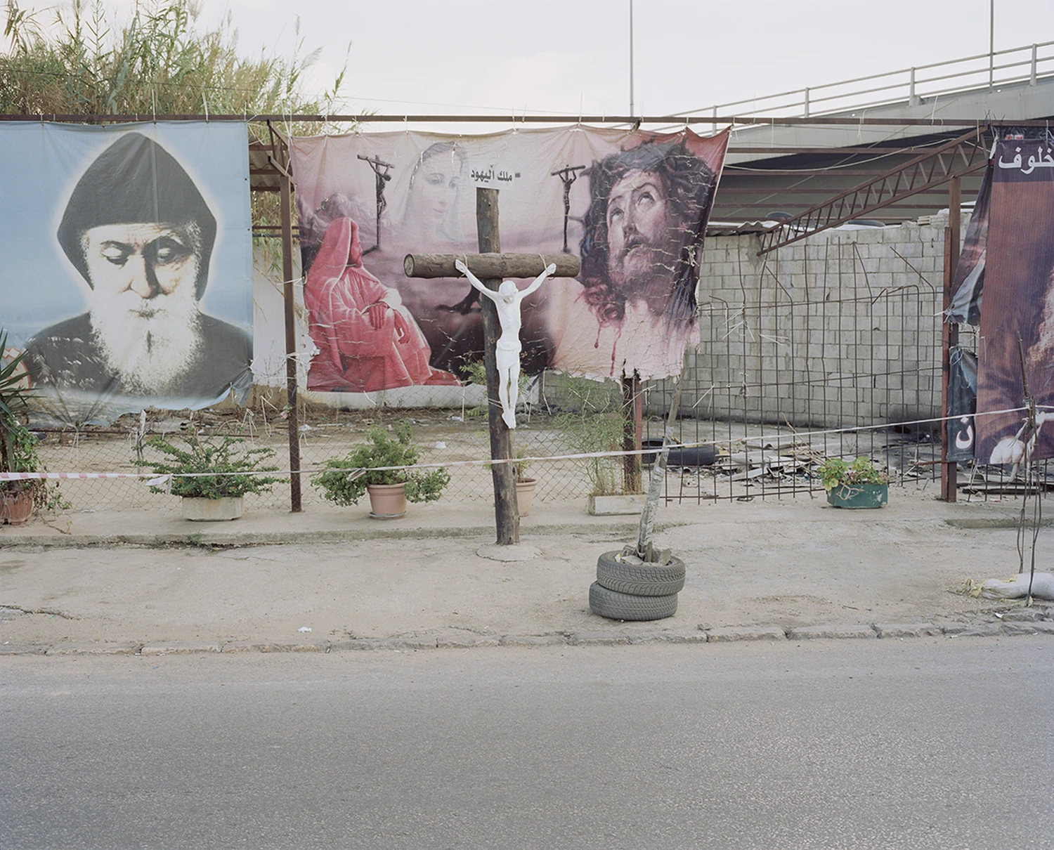 ASNAM (Idol Statues) by Georges Salameh — A white marble or plaster crucifix figure with outstretched arms occupies the center of a Lebanese street, mounted on a weathered concrete pedestal anchored...