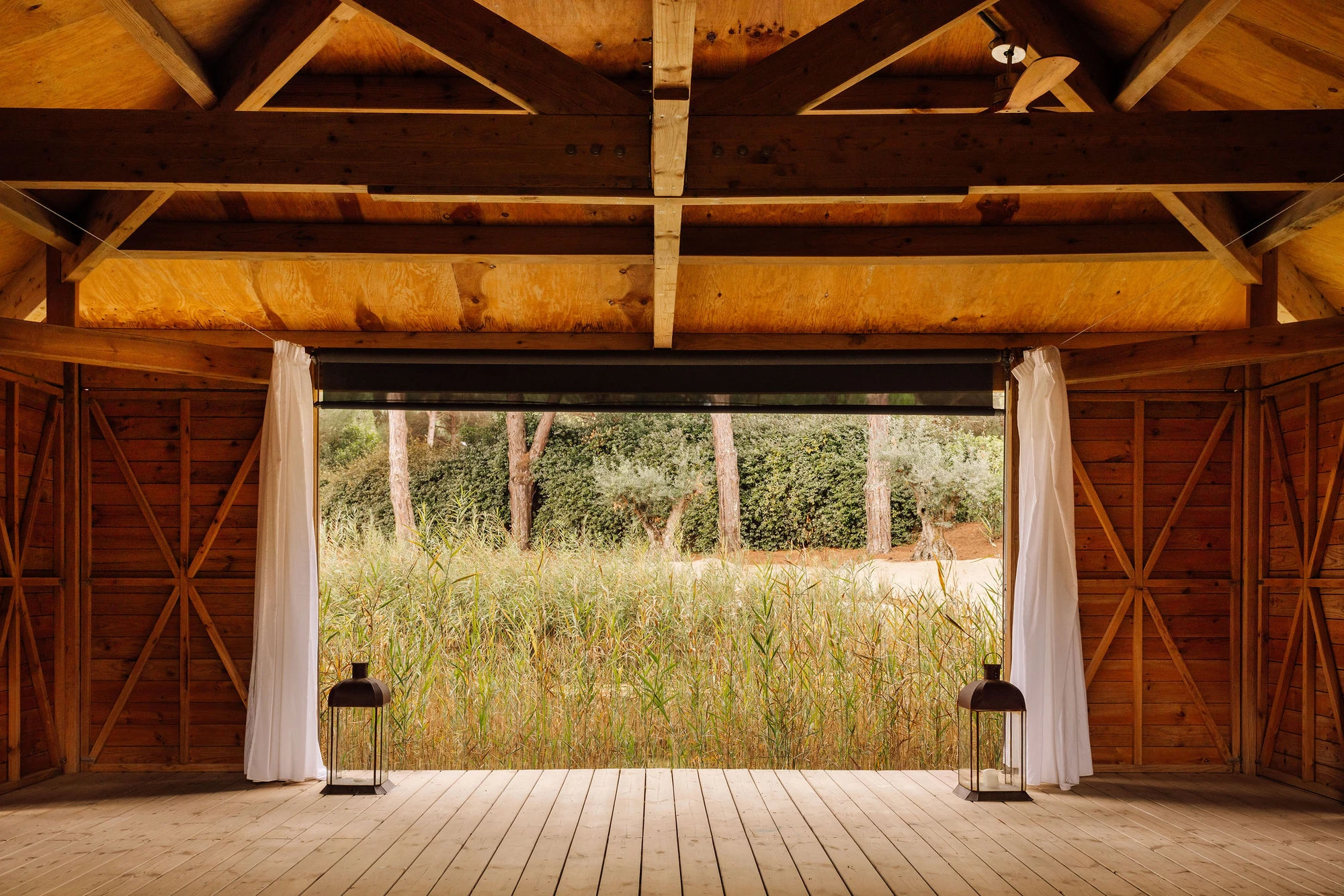Sublime Comporta — A timber pavilion interior frames a landscape vista through full-height glazed walls, with white linen curtains suspended from metal rods flanking the opening.