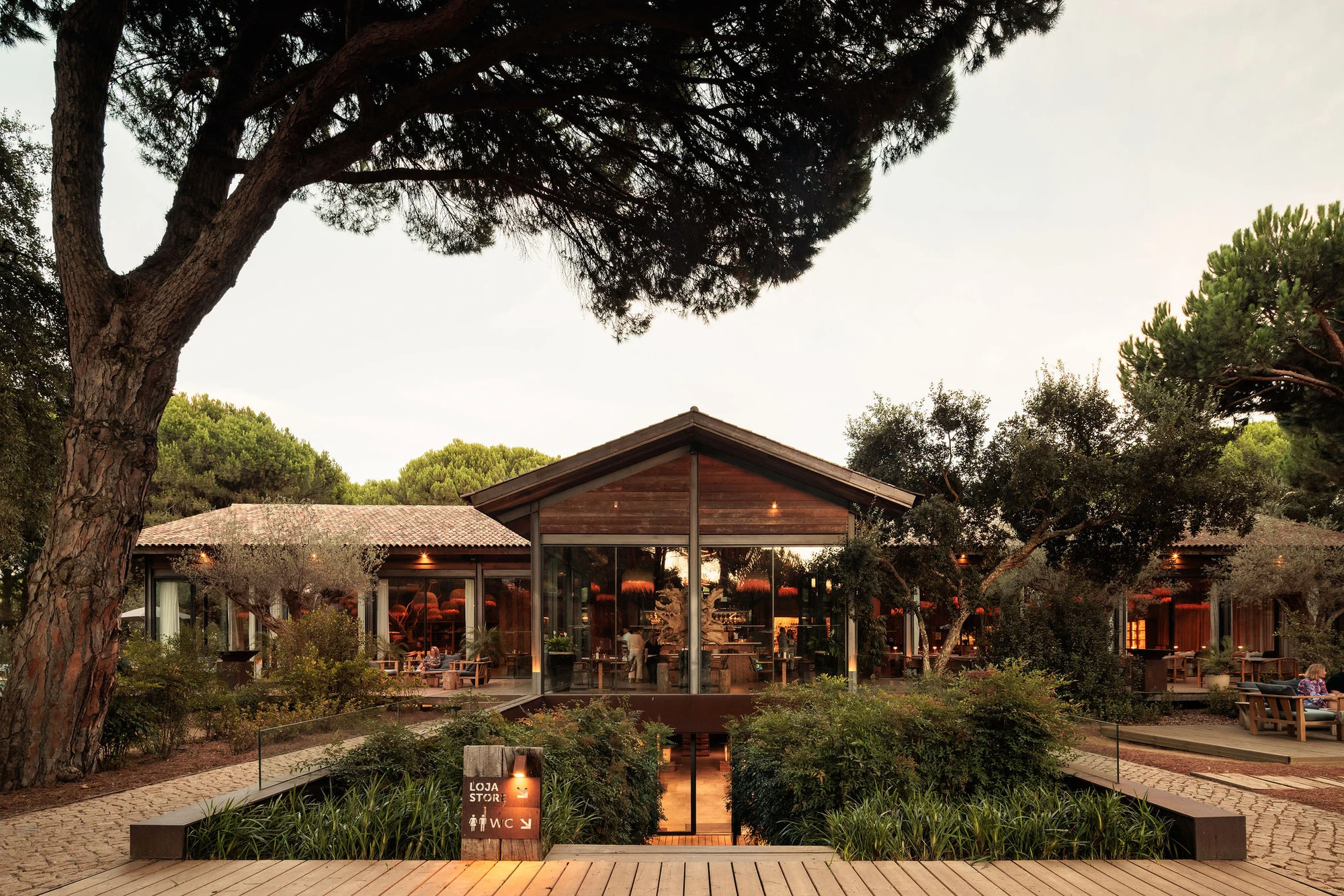 Sublime Comporta — A timber-clad pavilion with pitched roof and deep overhangs frames the entrance to Sublime Comporta, photographed at dusk.