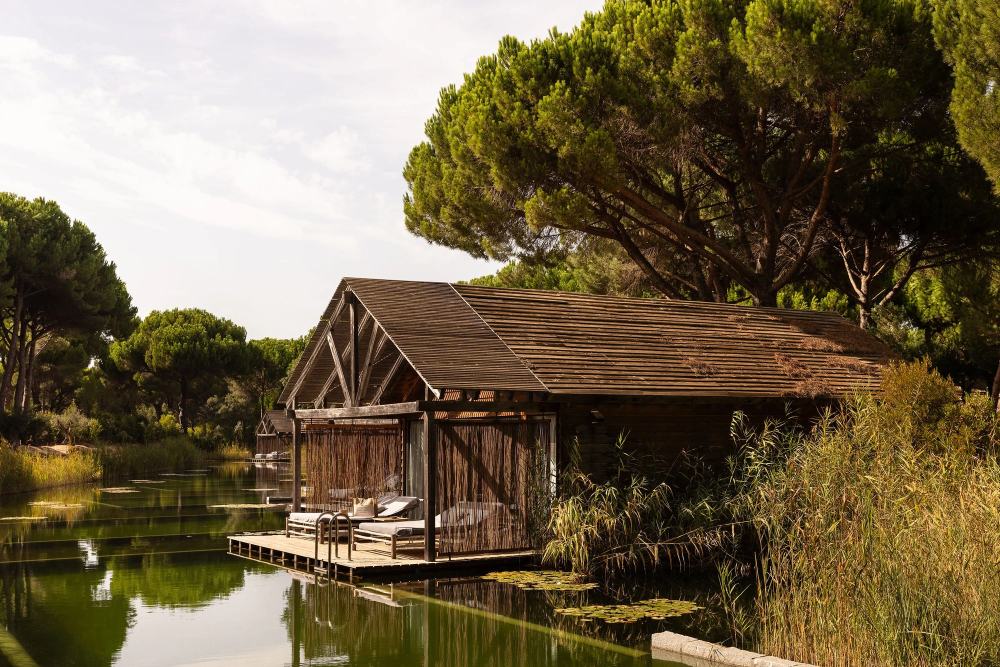 Sublime Comporta — A timber-clad pavilion with pitched roof and deep overhangs sits at the water's edge, accessed via a timber boardwalk that extends into the canal.