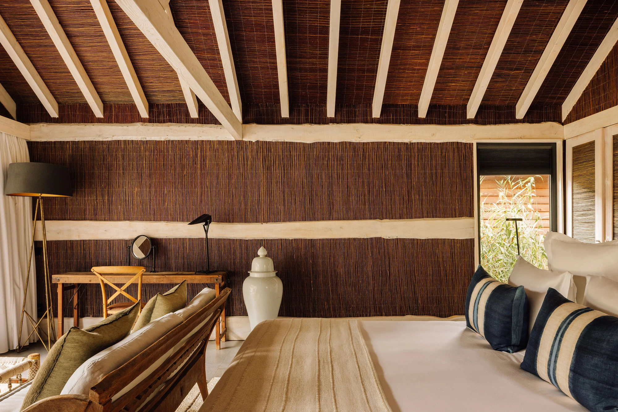 Sublime Comporta — A pavilion bedroom interior with exposed timber ceiling structure: cream-painted rafters cantilever over dark-stained reed or rattan wall paneling...