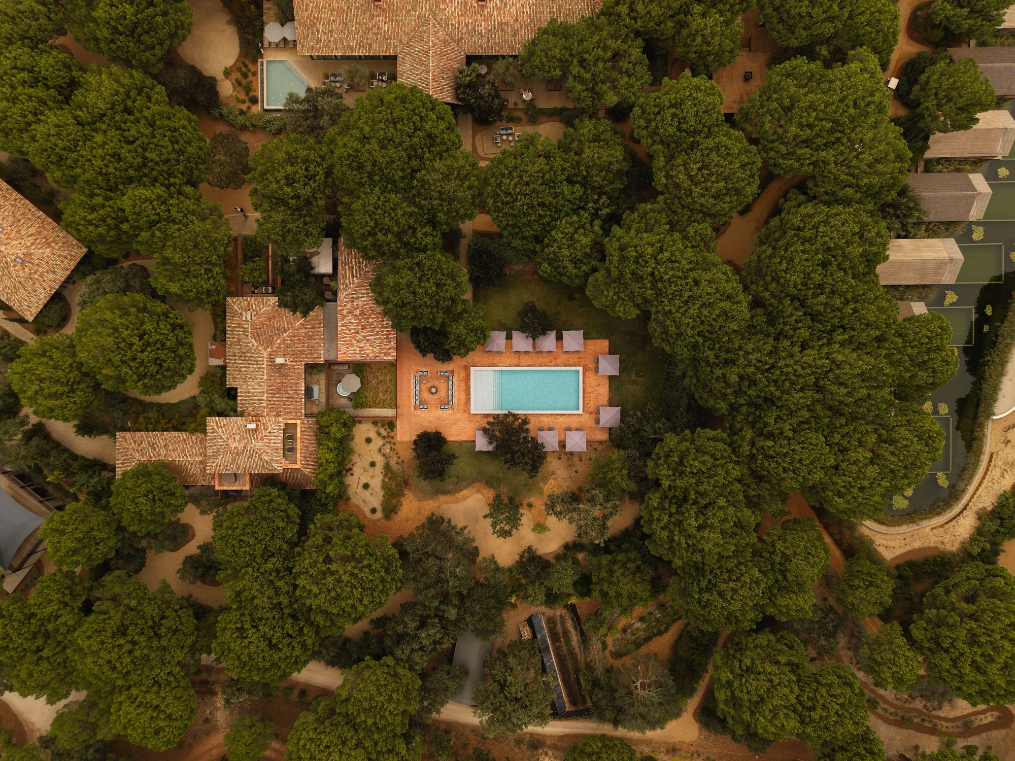 Sublime Comporta — Aerial view of a brick pavilion with terracotta-tiled pitched roof and rectangular swimming pool, surrounded by mature pine forest.
