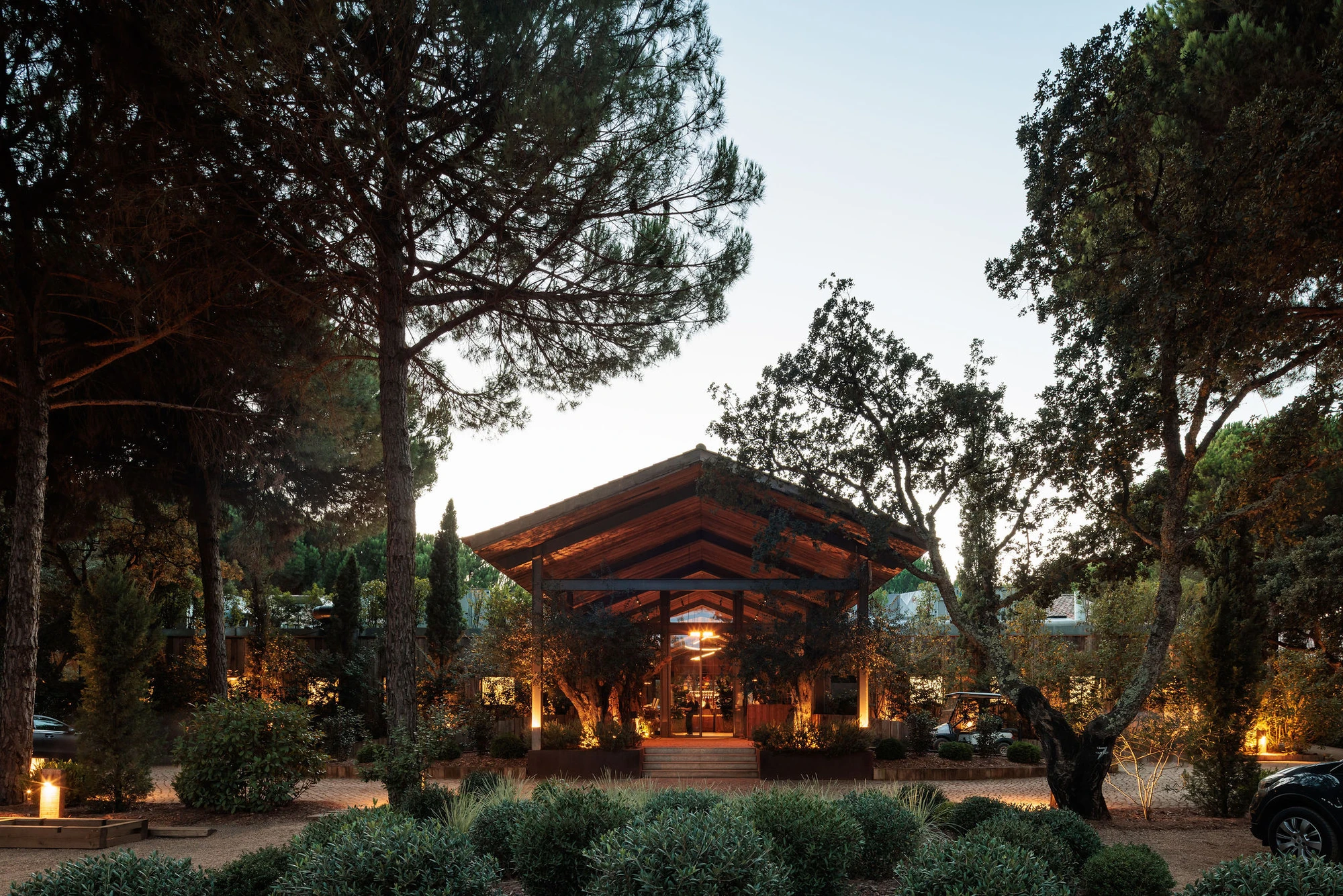 Sublime Comporta — A timber-clad pavilion with pitched roof and deep overhangs anchors a pine-forested clearing at dusk.
