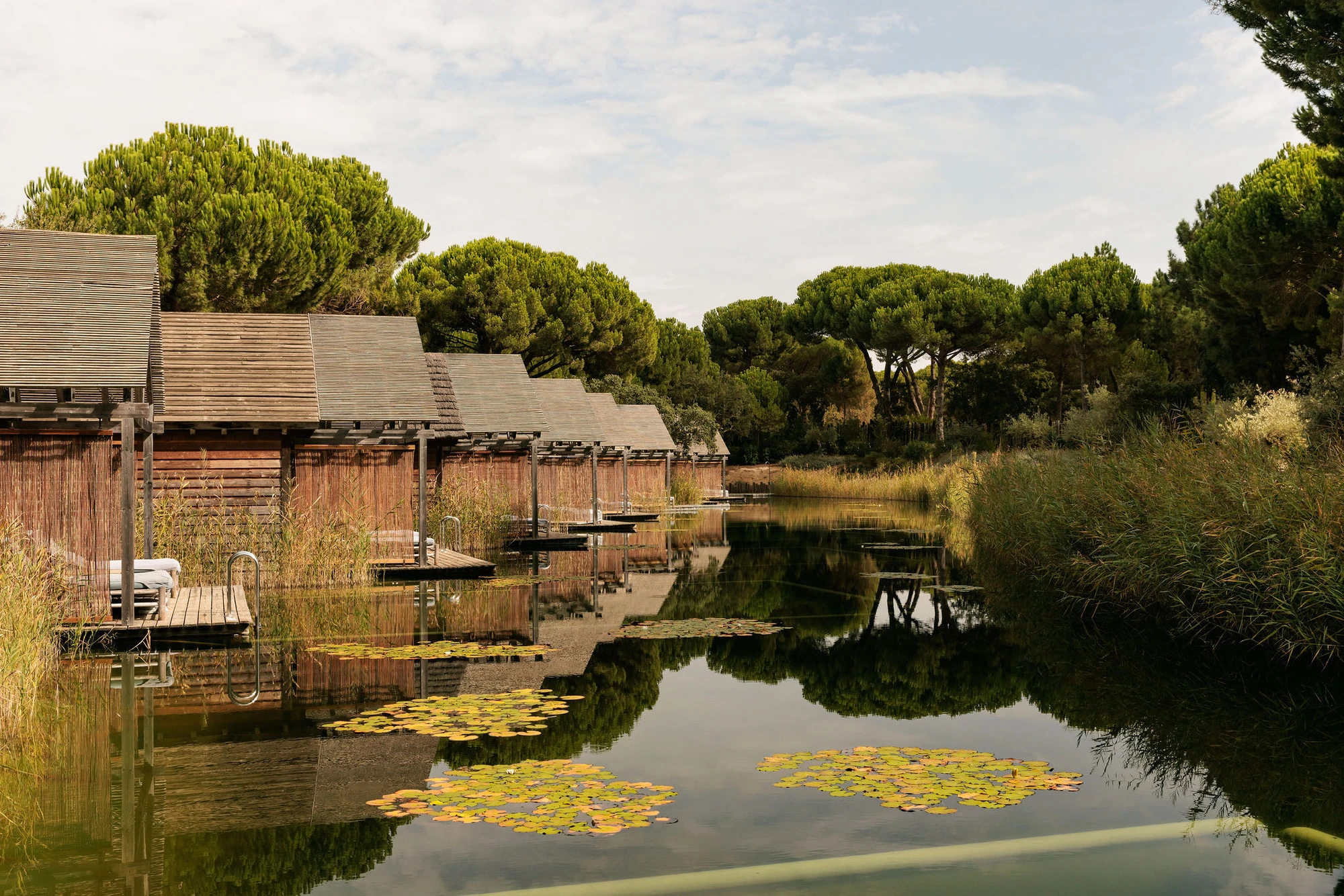 Sublime Comporta — Timber-clad pavilions with pitched roofs and deep overhangs line a still waterway, their weathered wooden facades reflected in the glassy surface below.