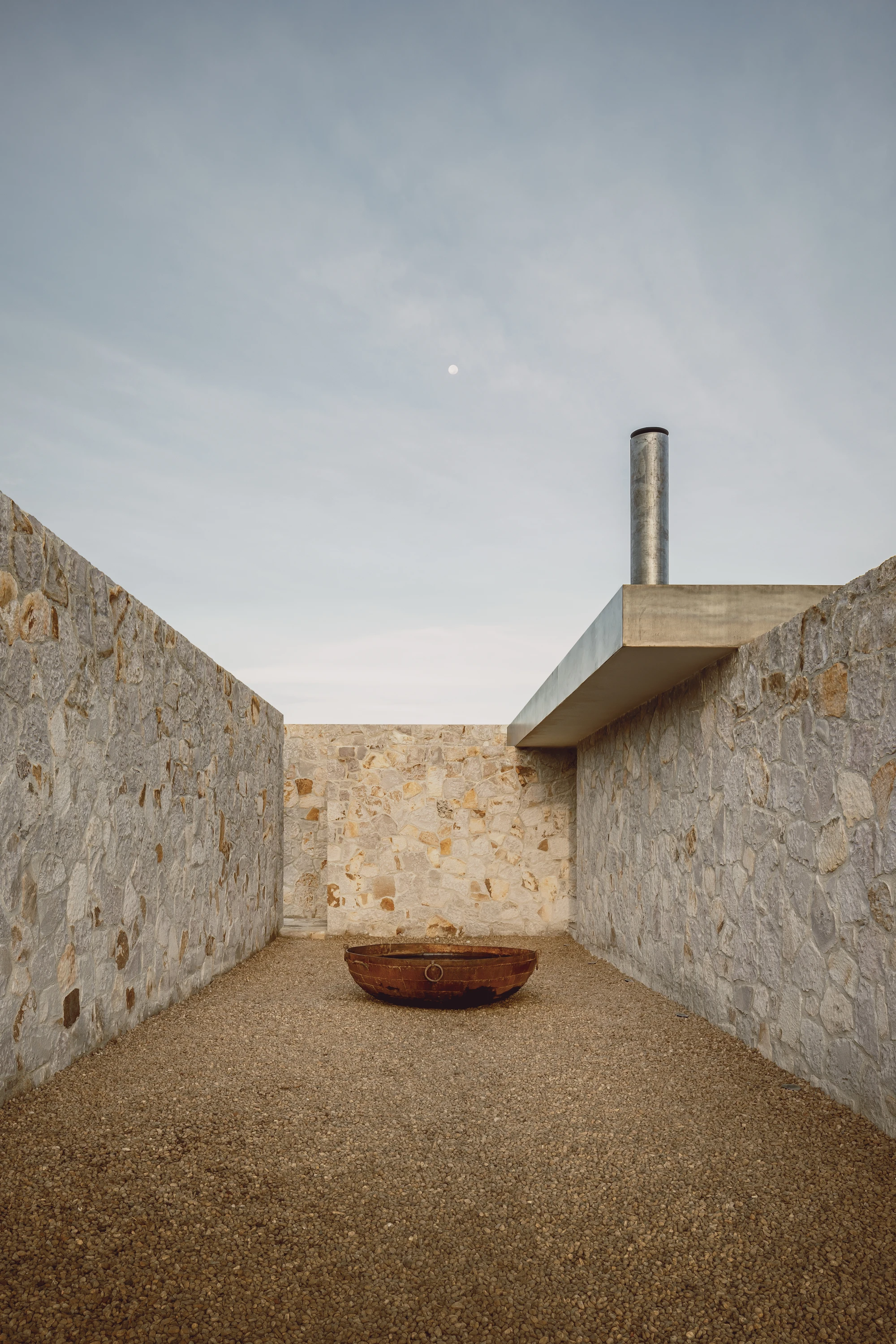 Enso House II by HW Studio — A narrow passage framed by coursed stone walls with horizontal stratification in cream and ochre tones creates a compressed perspective toward a monolithic...