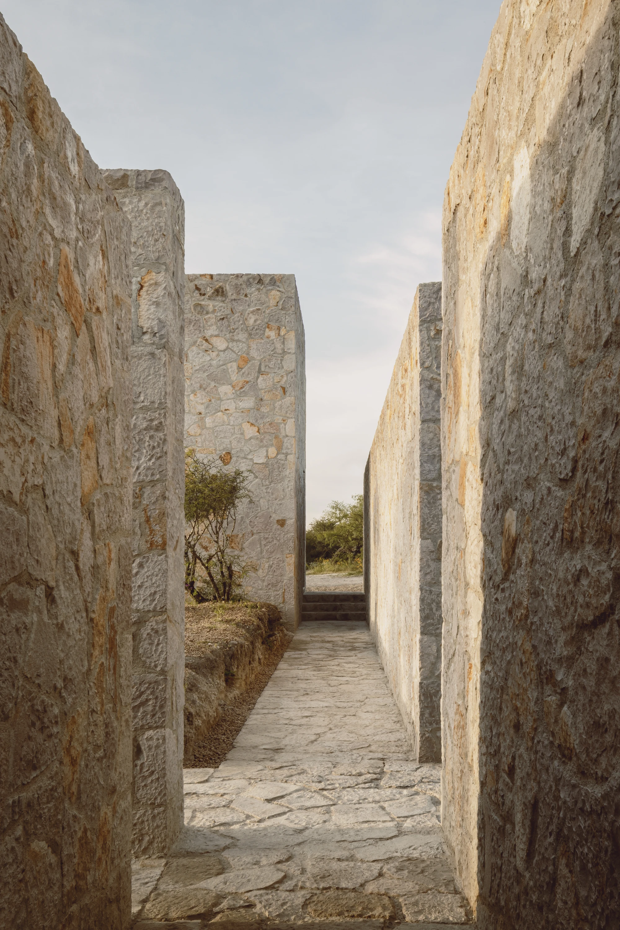 Enso House II by HW Studio — A narrow stone corridor frames the landscape beyond, its coursed walls constructed from pale ochre and grey stone with visible horizontal stratification.