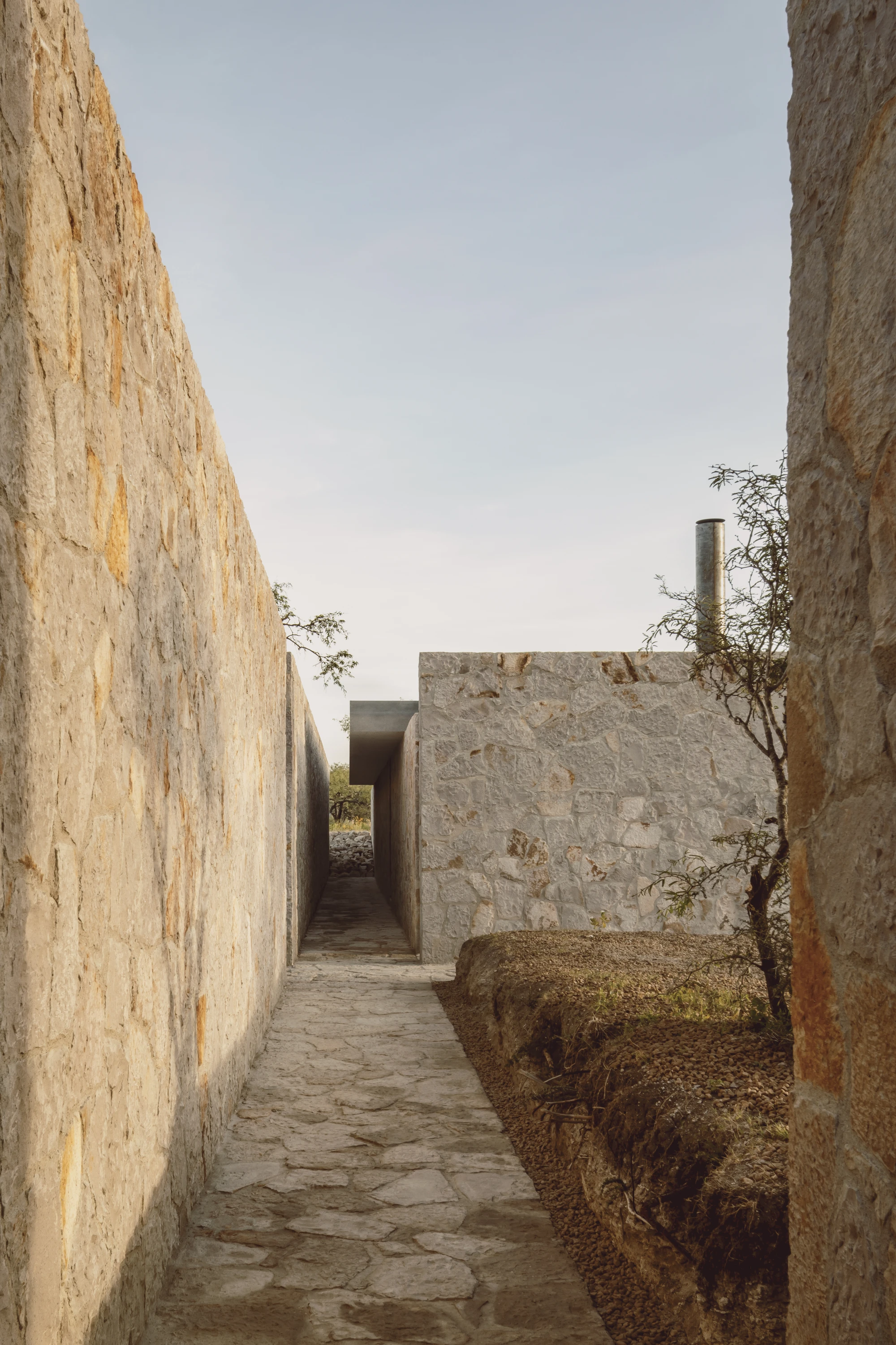 Enso House II by HW Studio — A narrow stone passage frames the residential structure beyond, its coursed masonry walls exhibiting horizontal geological stratification in cream and ochre...