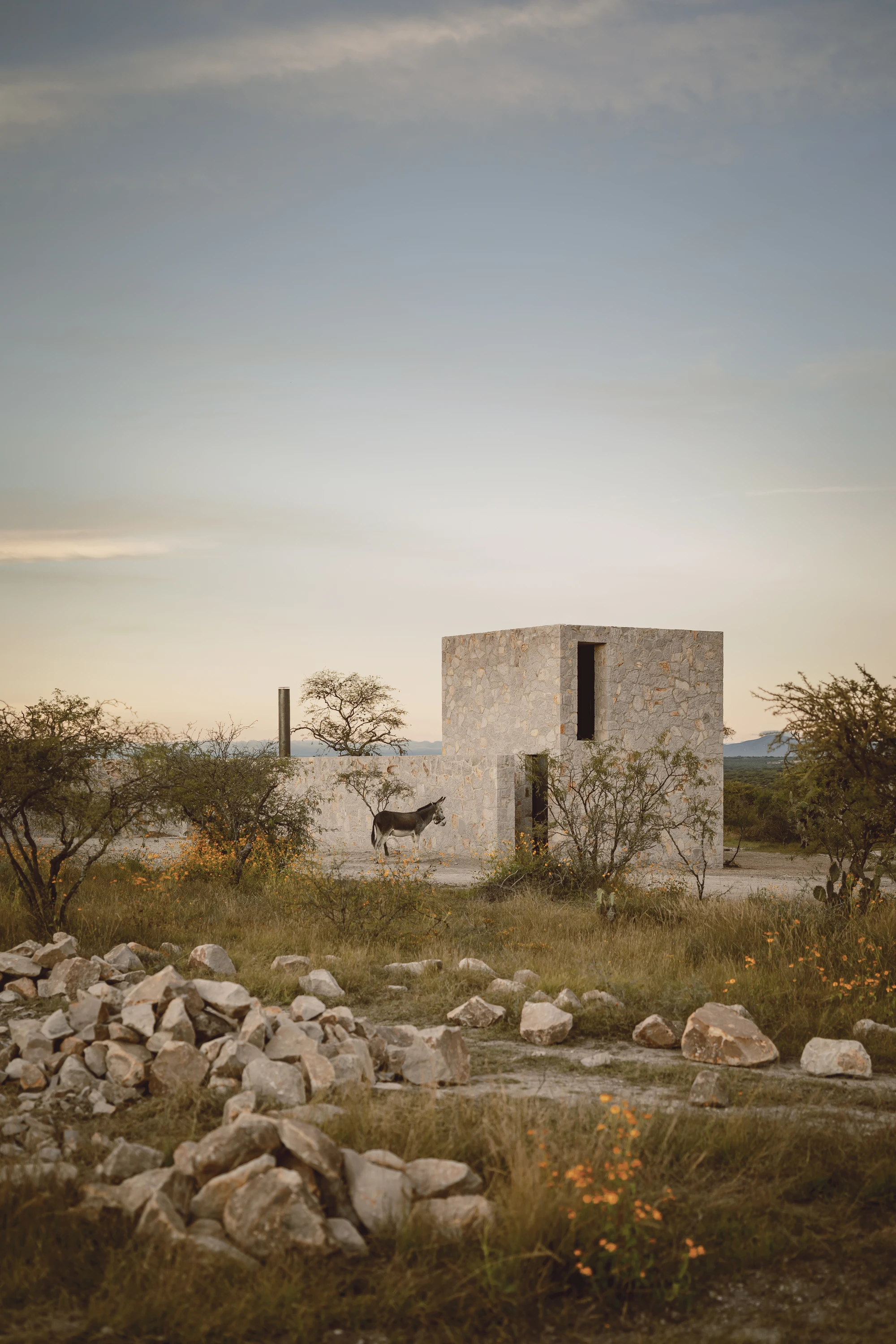 Enso House II by HW Studio — A coursed stone structure with horizontal stratification rises from the mesquite-dotted landscape, its rectangular window opening cut cleanly through the...
