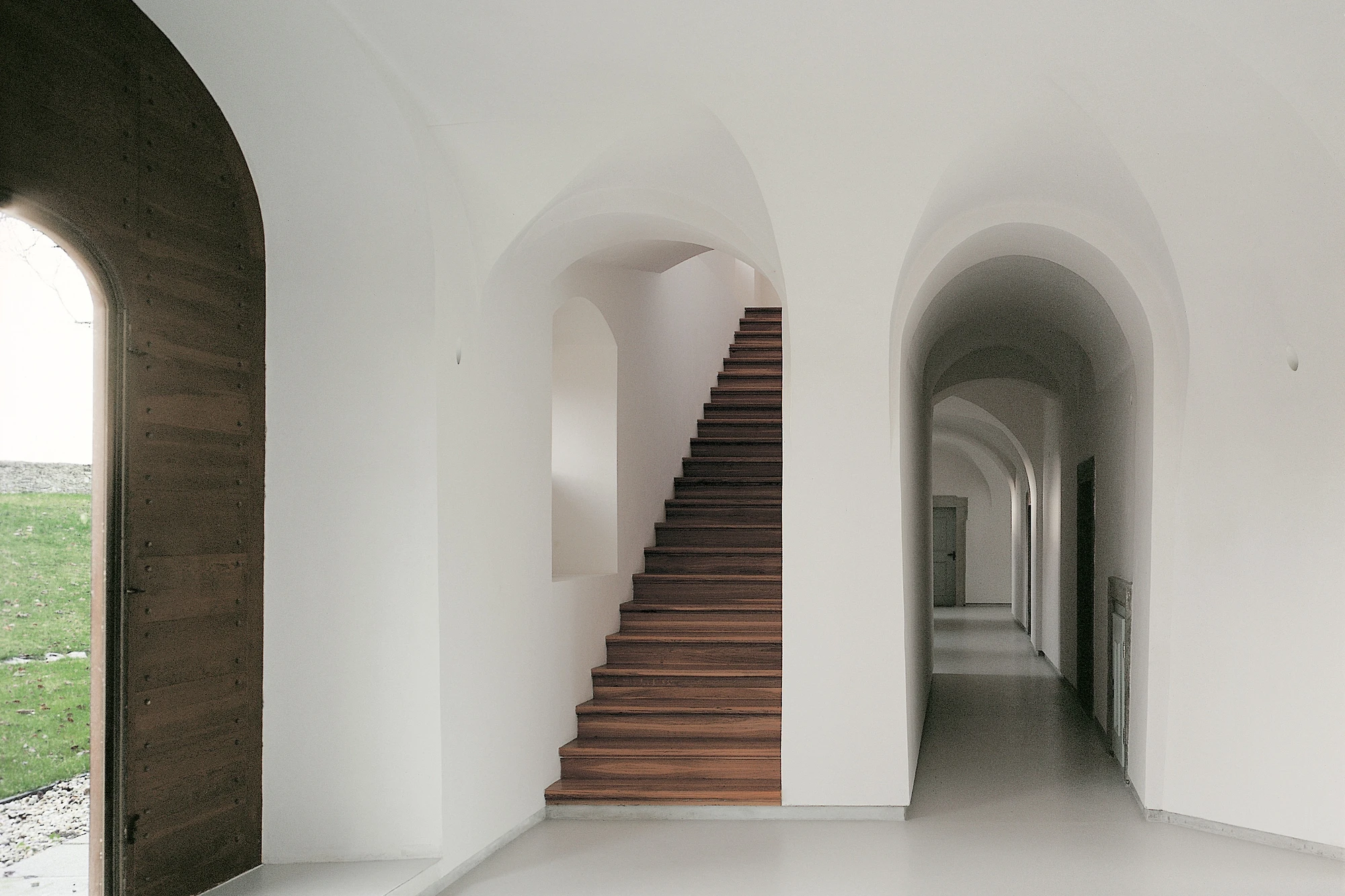 Abbey of Our Lady of Nový Dvůr by John Pawson — A vaulted corridor lined with repeated barrel-vault arches in white plaster frames a wooden staircase ascending through the center.