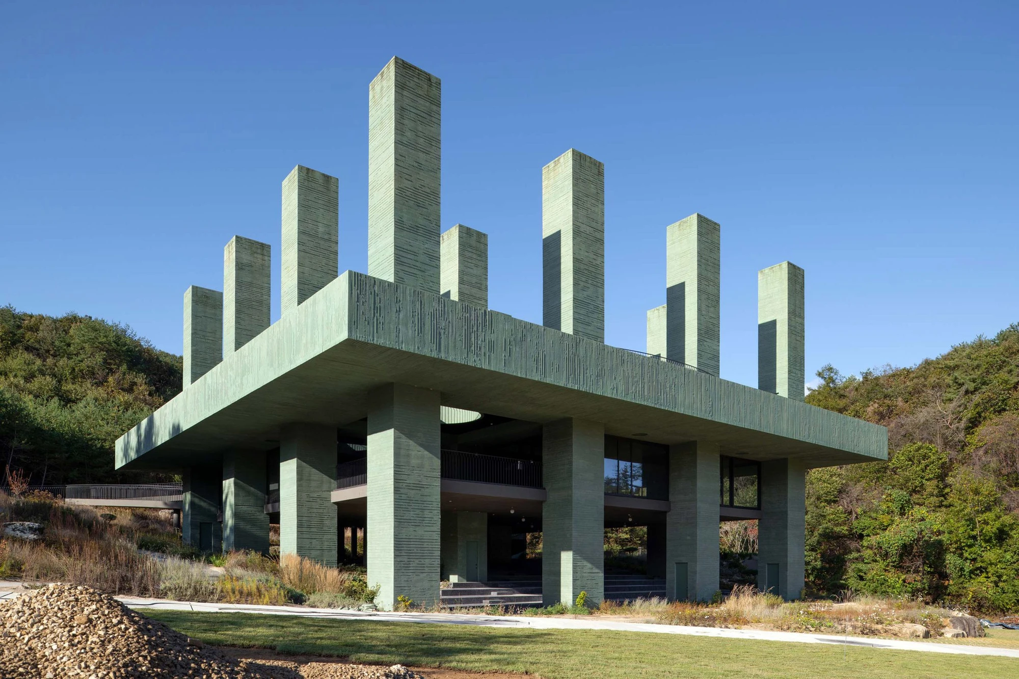 REST Garden Restaurant by Pezo von Ellrichshausen — Nine rectangular concrete columns arranged in a strict grid rise from a cantilevered platform, their surfaces clad in soft sage-tinted concrete with dark...