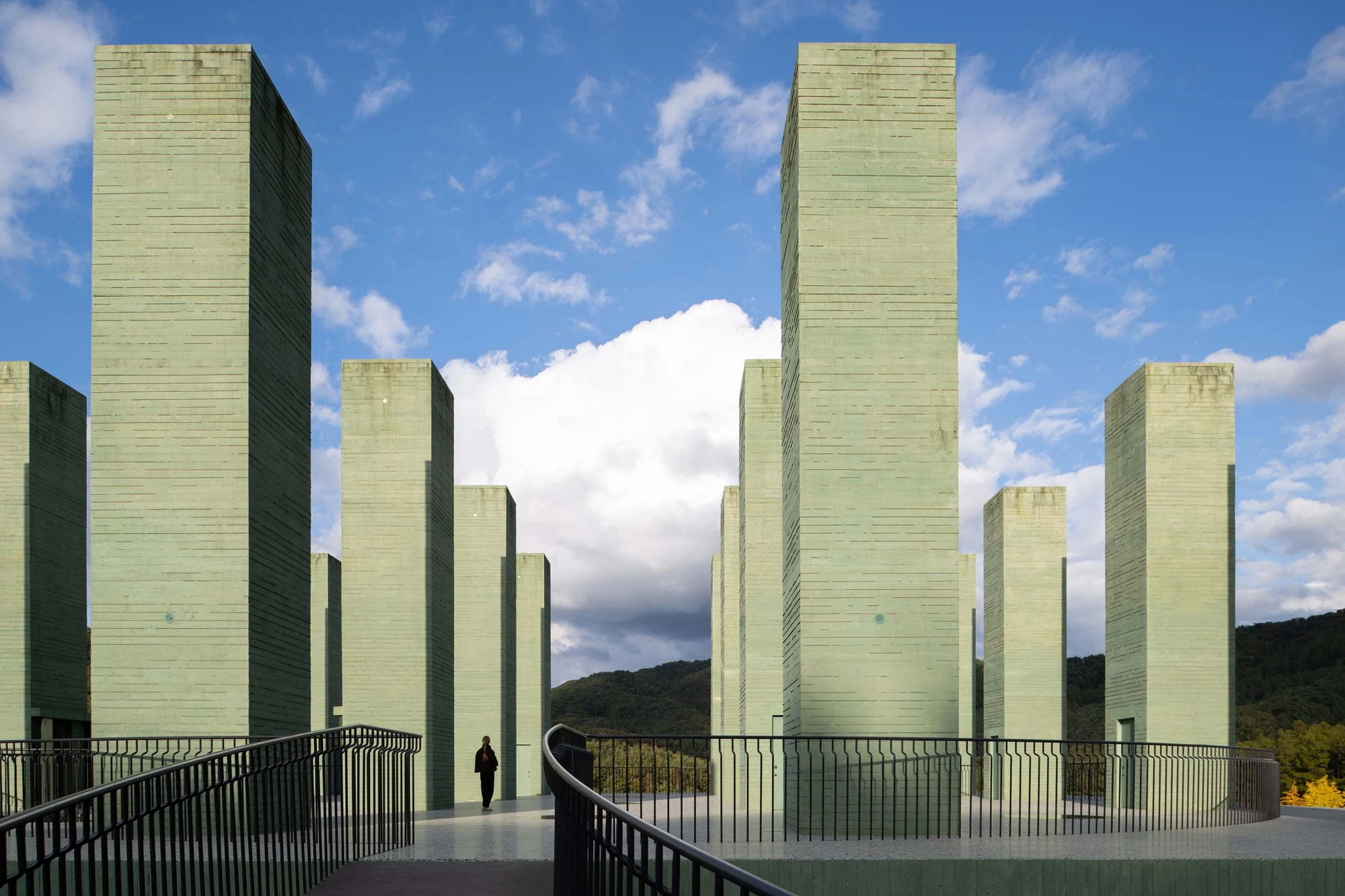 REST Garden Restaurant by Pezo von Ellrichshausen — Nine vertical concrete columns in sage-tinted cast material dominate the composition, arranged in a strict grid across an elevated platform cantilevered...