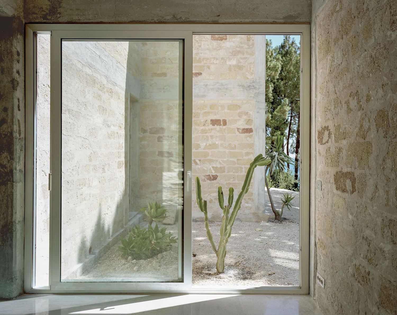 Guardia di Finanza by Brandlhuber+ — A threshold view through paired glass doors with dark aluminum frames, opening from a concrete-floored interior onto a gravel courtyard.