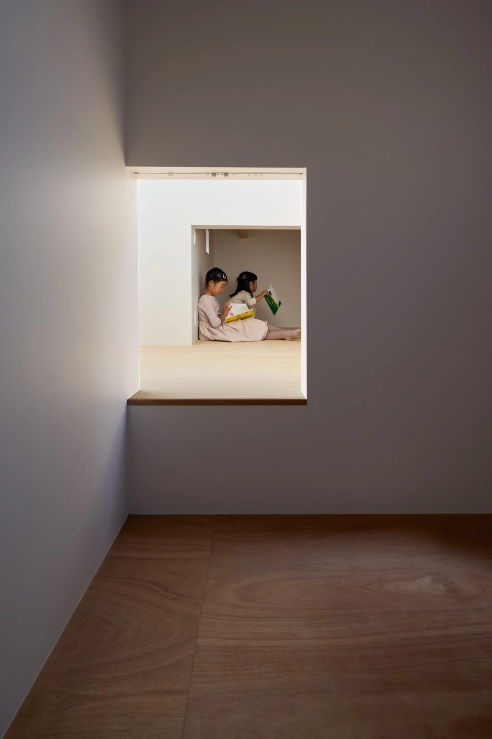House in Hirano by Fujiwaramuro Architects — Two children sit cross-legged within a recessed alcove carved into the building's interior, framed by white render walls and a timber threshold.