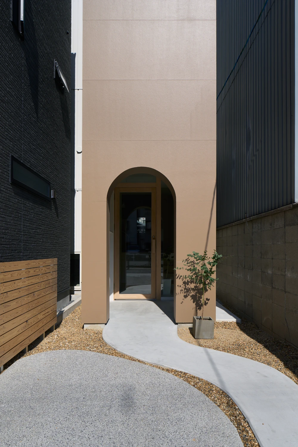 House in Hirano by Fujiwaramuro Architects — A narrow passage between two buildings converges toward a monolithic peach-render facade punctuated by a single arched dark doorway.