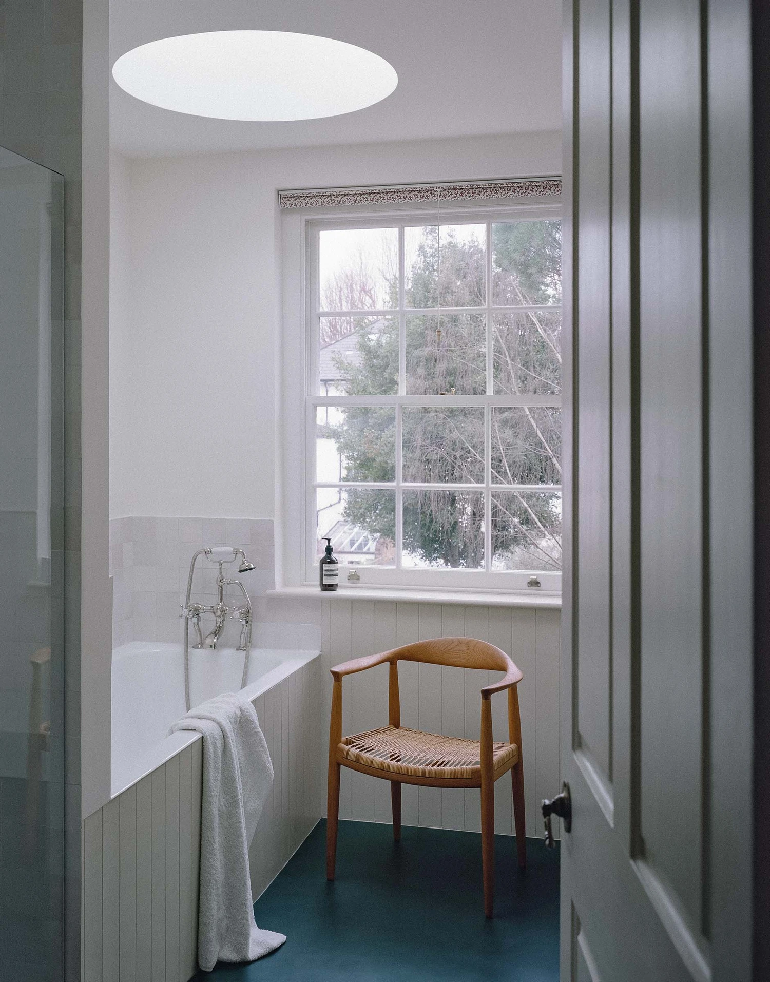 Barnsbury House by Architecture for London — A bathroom in Barnsbury House featuring a freestanding white bathtub positioned beneath a large multi-paned window framing bare winter trees.