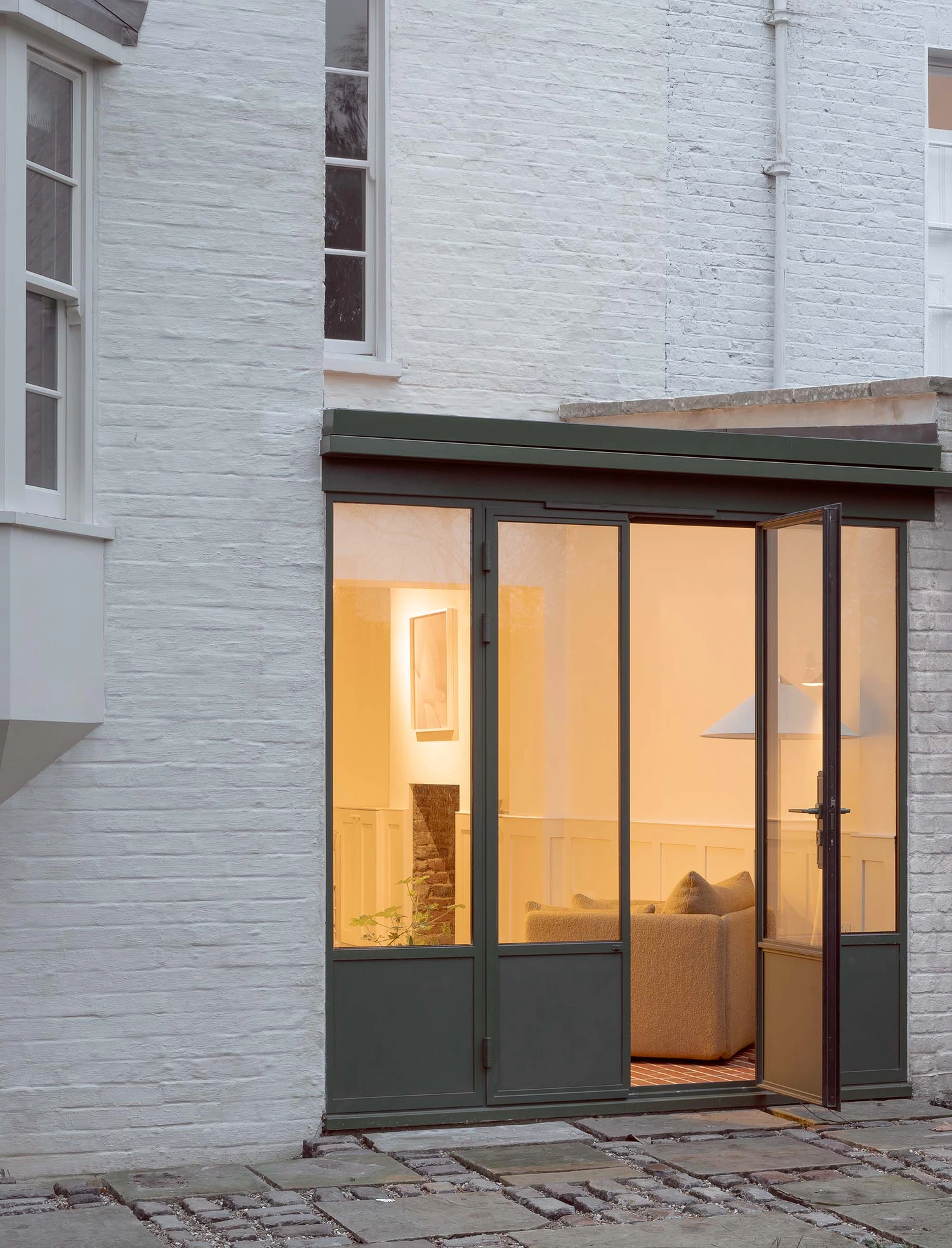 Barnsbury House by Architecture for London — A timber-framed garden room extension opens onto a paved courtyard through multi-paned glazed doors with dark-painted frames.