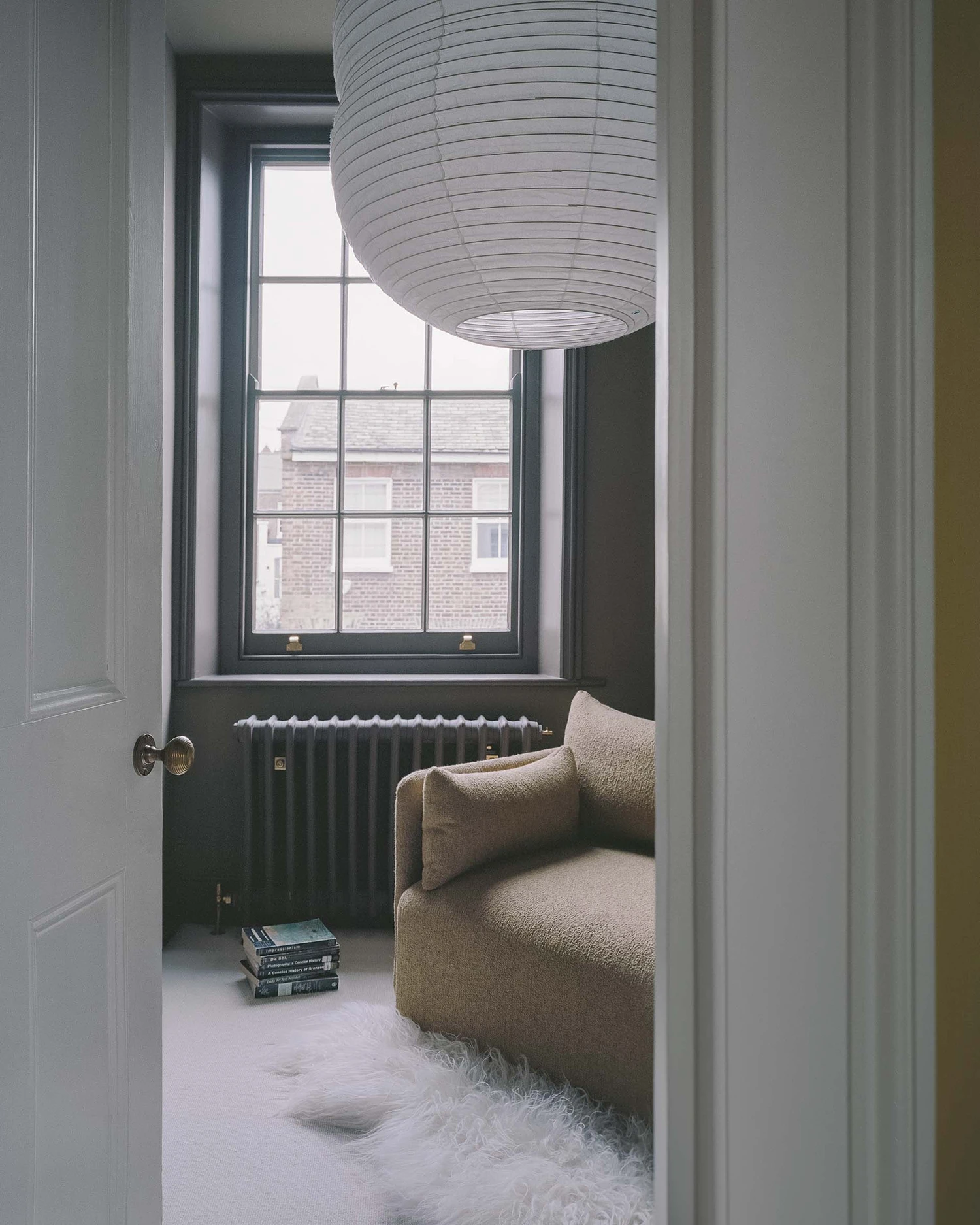 Barnsbury House by Architecture for London — A reading nook framed through an open doorway reveals a beige upholstered armchair positioned beneath a tall sash window with dark-painted frame and...
