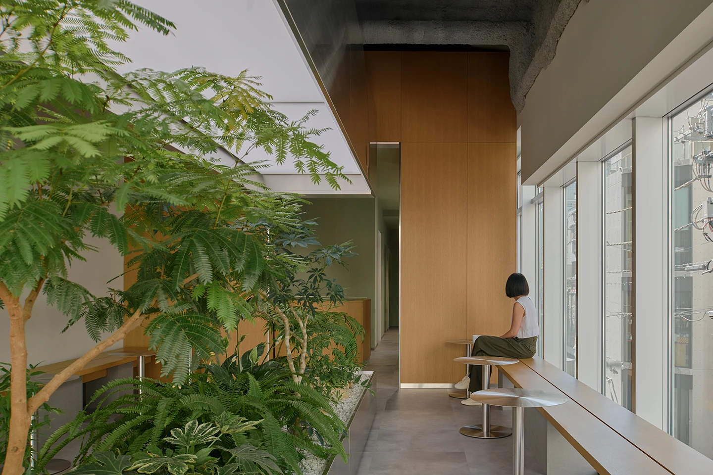 Alchemist Aoyama by Wynk Collaborative — A seated figure occupies a wooden bench integrated into the perimeter of Alchemist Aoyama's upper level, positioned as an observer of the dense planted...