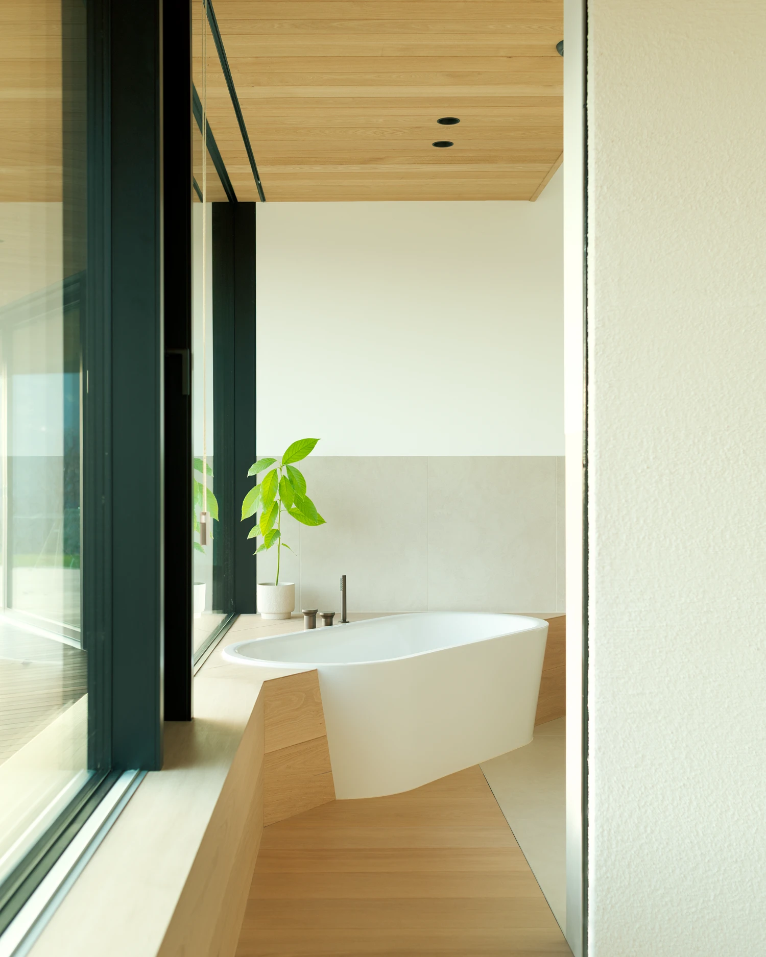 Casa Conte by Messner Architects — A freestanding white oval bathtub occupies a timber-floored bathroom, positioned parallel to a floor-to-ceiling glazed opening framed in black aluminum.