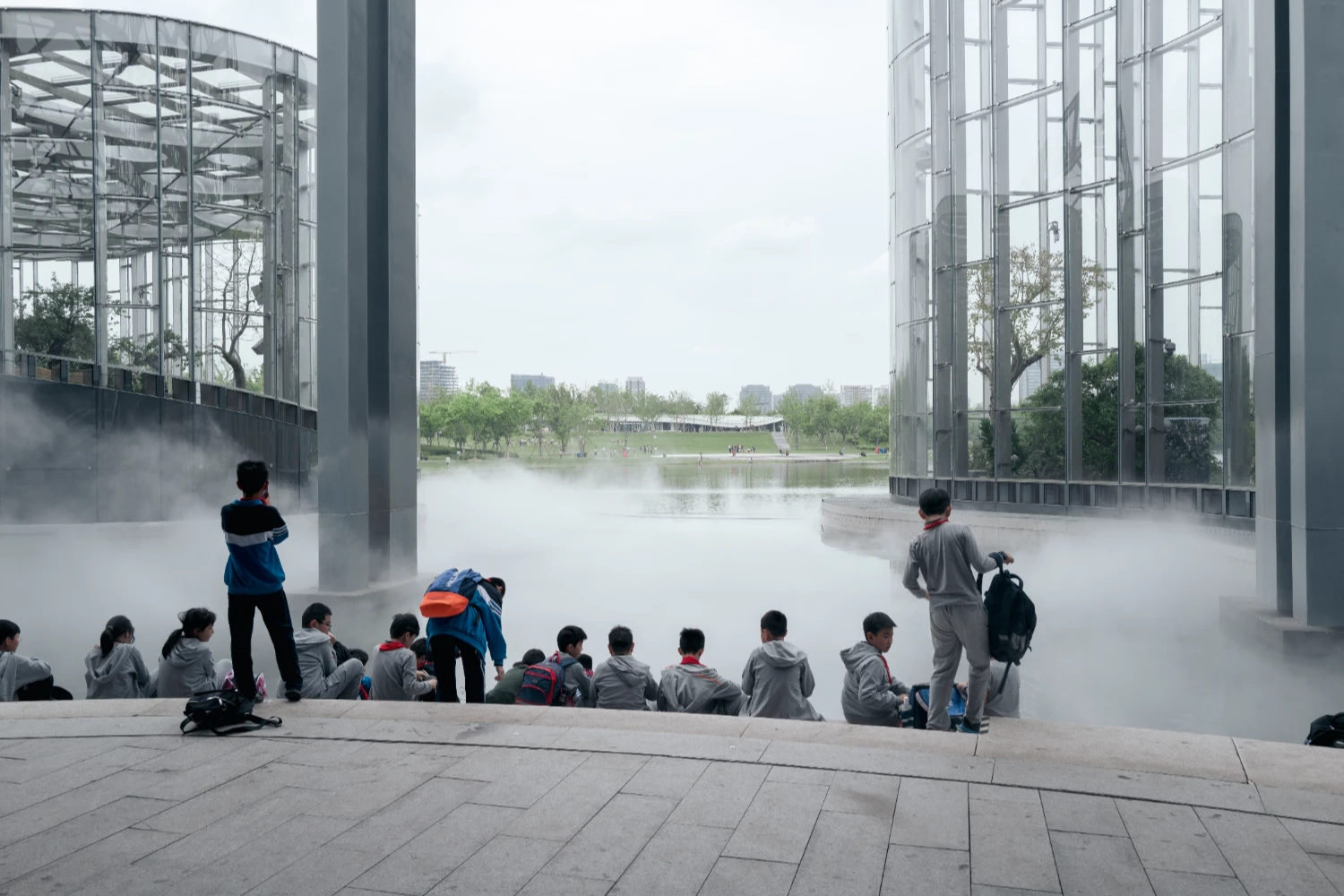 Expo Cultural Park Greenhouse Garden by Delugan Meissl captured by Marcus Yau — Children sit in rows on concrete paving facing a threshold of artificial fog that obscures the glazed greenhouse interior.