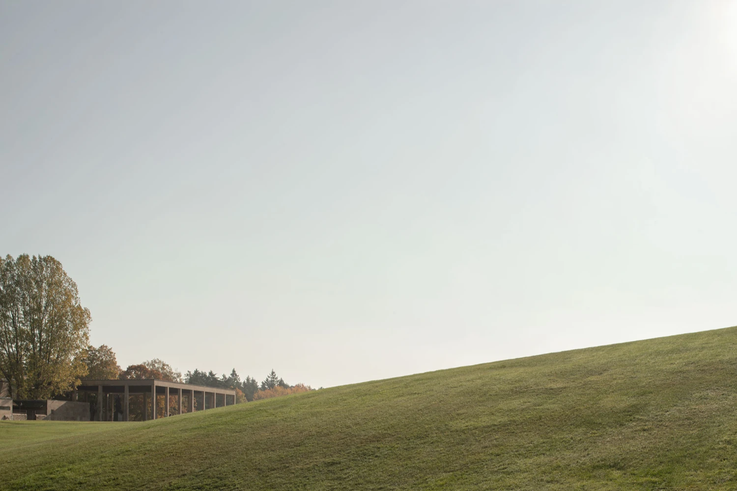 Woodland Cemetery Stockholm captured by Andy Liffner — A modernist timber structure emerges from the forest edge, its post-and-beam construction defining a linear pavilion against the sloping Nordic landscape.