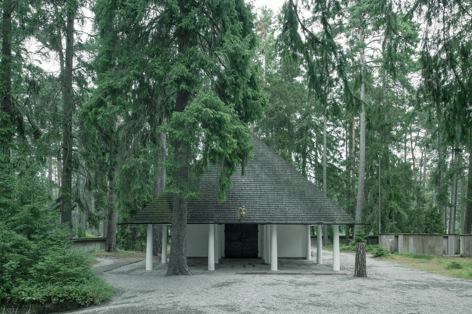 Woodland Cemetery Stockholm captured by Andy Liffner — A modernist pavilion emerges within dense Nordic forest, its pitched roof clad in dark-grey tile ascending to a triangular apex.