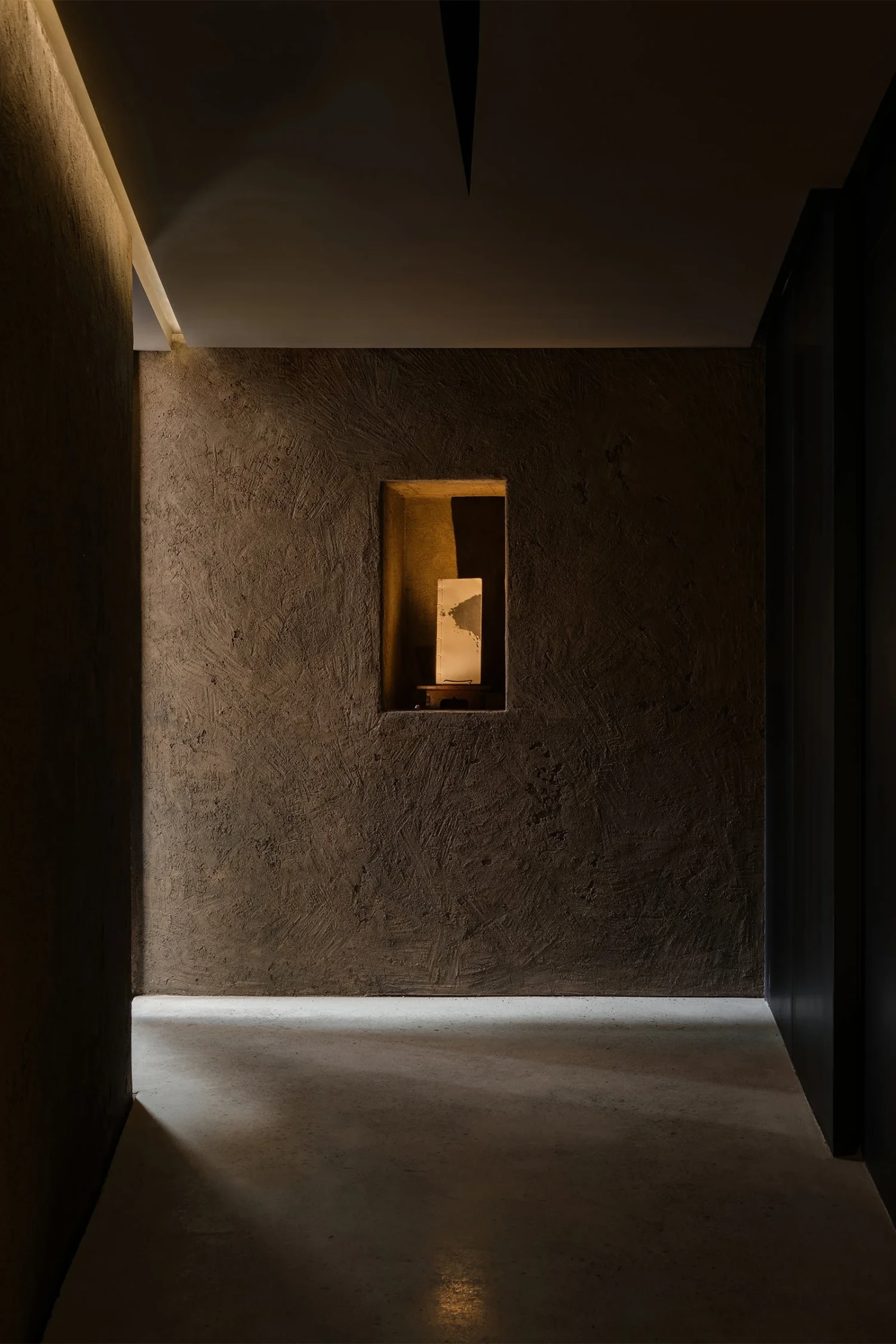 The Graphic bookstore by OONN Metaworks + U.LAB — A concrete corridor frames a distant wooden doorway, its warm ochre tone intensified by the surrounding shadow.