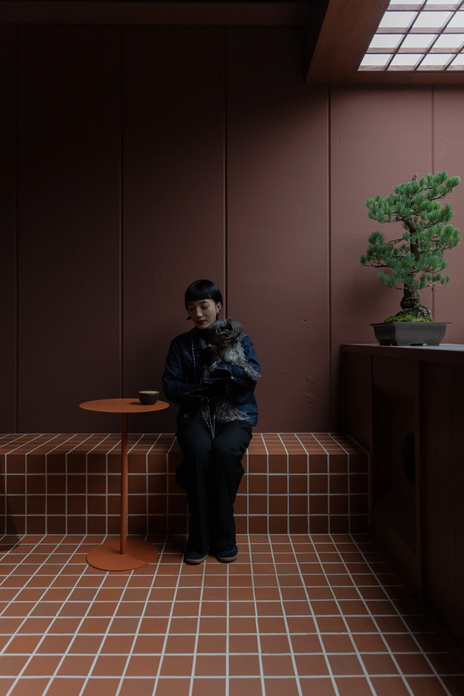 uru coffee by TRANSFORM — A figure in dark navy jacket sits centered on a terracotta-tiled bench, holding a small gray dog.