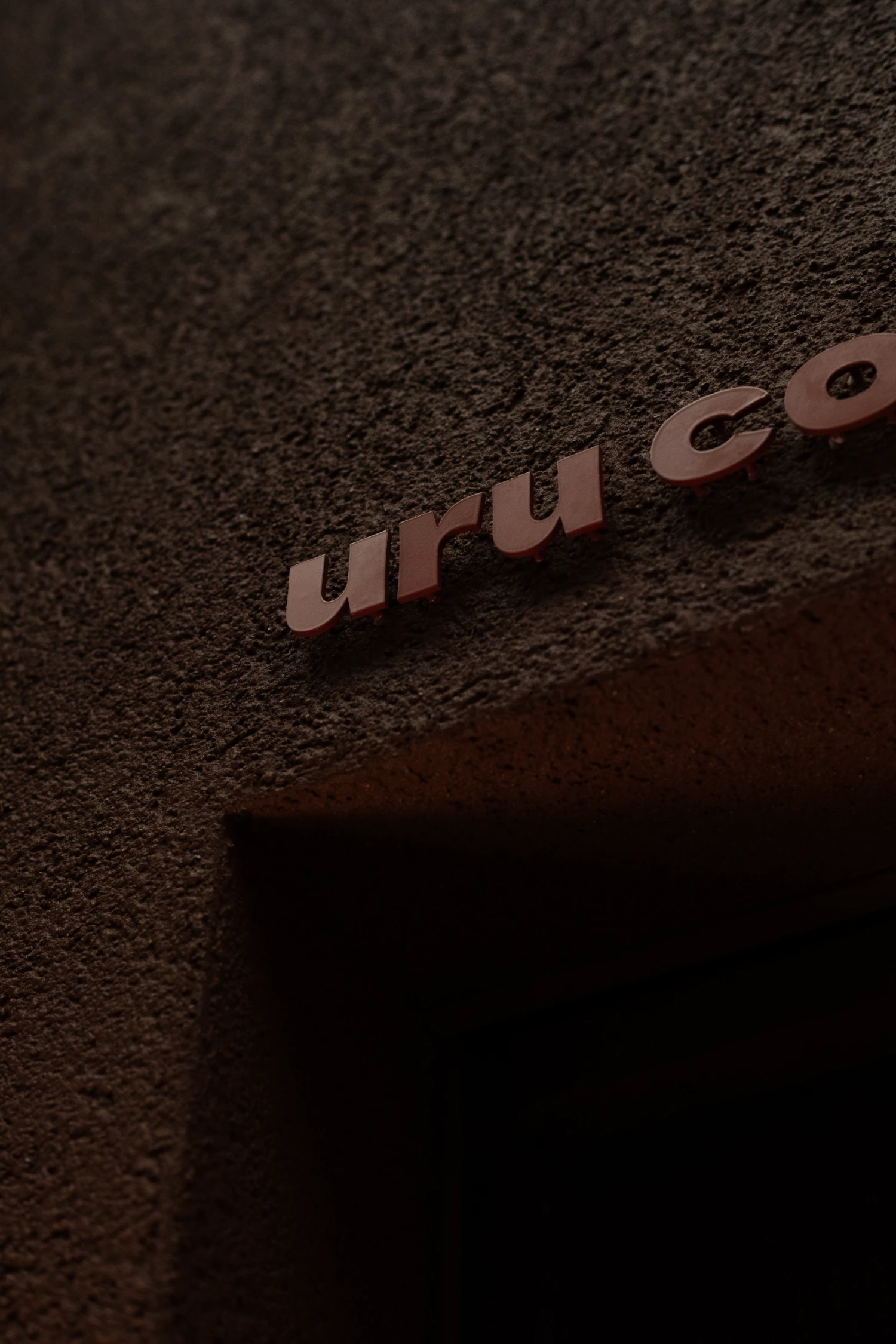 uru coffee by TRANSFORM — A close-up detail of the uru coffee signage mounted on a textured terracotta wall, captured in dramatic side-lighting that emphasizes the dimensional...