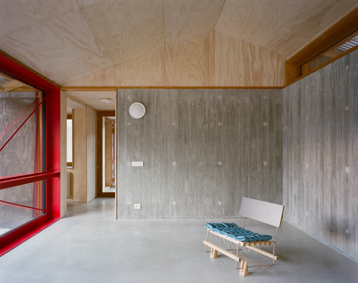 Casa BM by ErranteArchitetture — An unfinished interior volume defined by board-formed concrete walls and a plywood ceiling, punctuated by timber structural beams and a warm wood-framed...