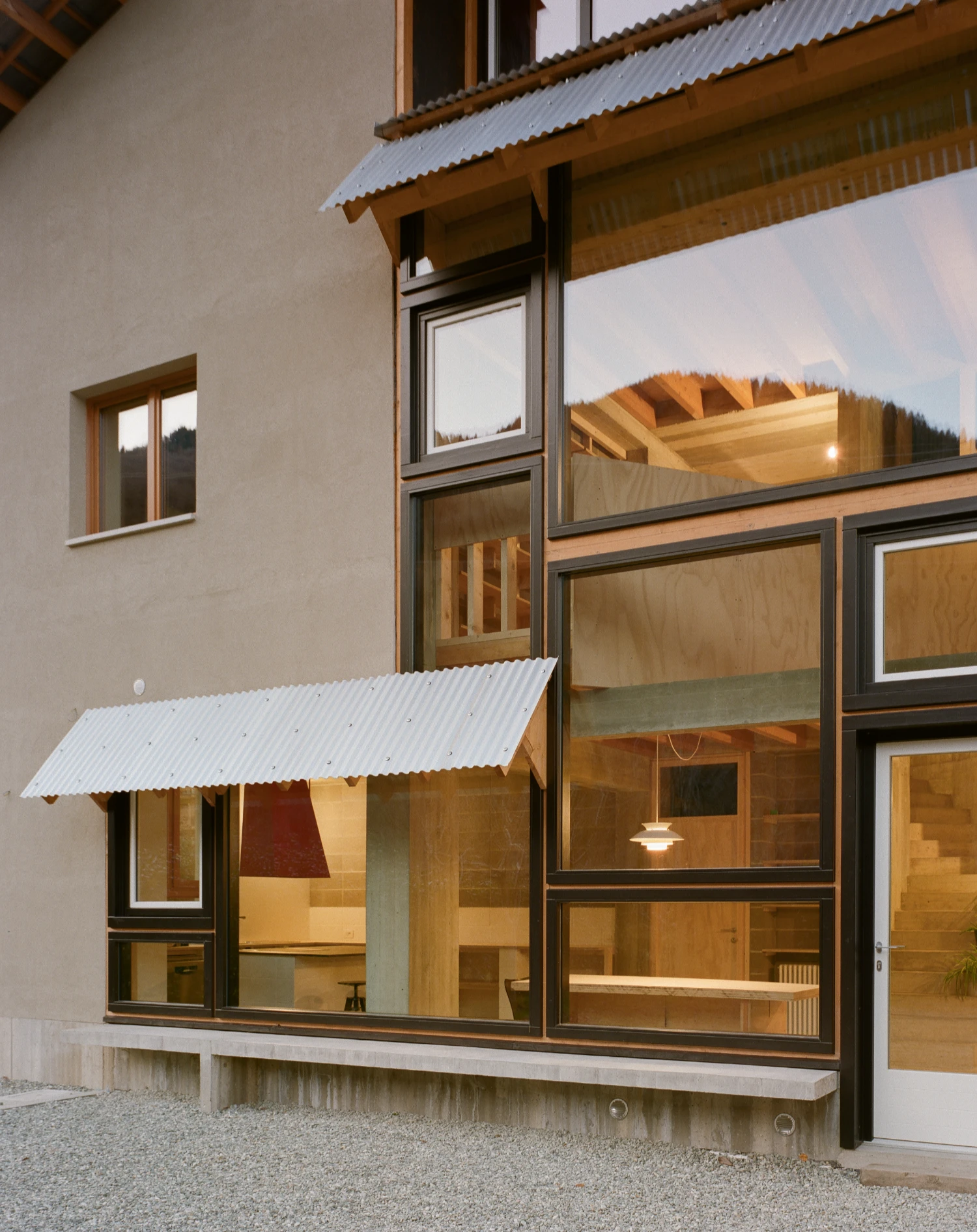 Casa BM by ErranteArchitetture — The garden facade reveals Casa BM's strategic intervention: a timber-frame pavilion with deep-pitched corrugated metal roof extends from the rendered...