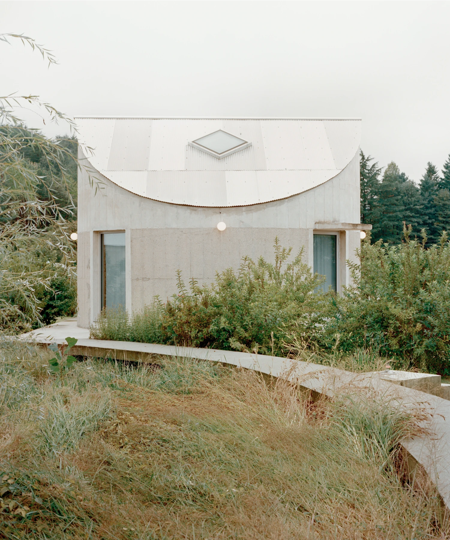 Hoji Gangneung Houses by AOA Architects captured by Danny Kai — A rendered concrete pavilion with a distinctive curved roof ridge and octagonal footprint emerges from an overgrown landscape of self-seeded grasses and...