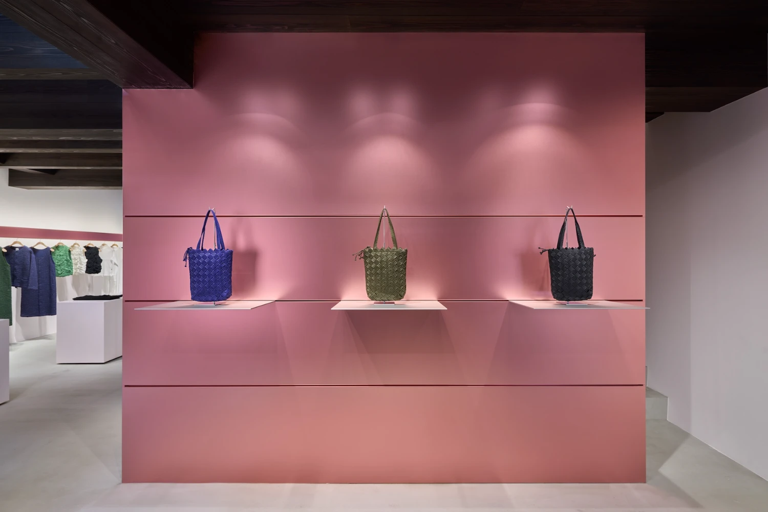 Issey Miyake HaaT flagship in Kyoto by Tokujin Yoshioka — Interior view of the HaaT flagship's display wall, a monolithic pink-rendered partition housing three woven cylindrical bags in blue, olive, and charcoal.