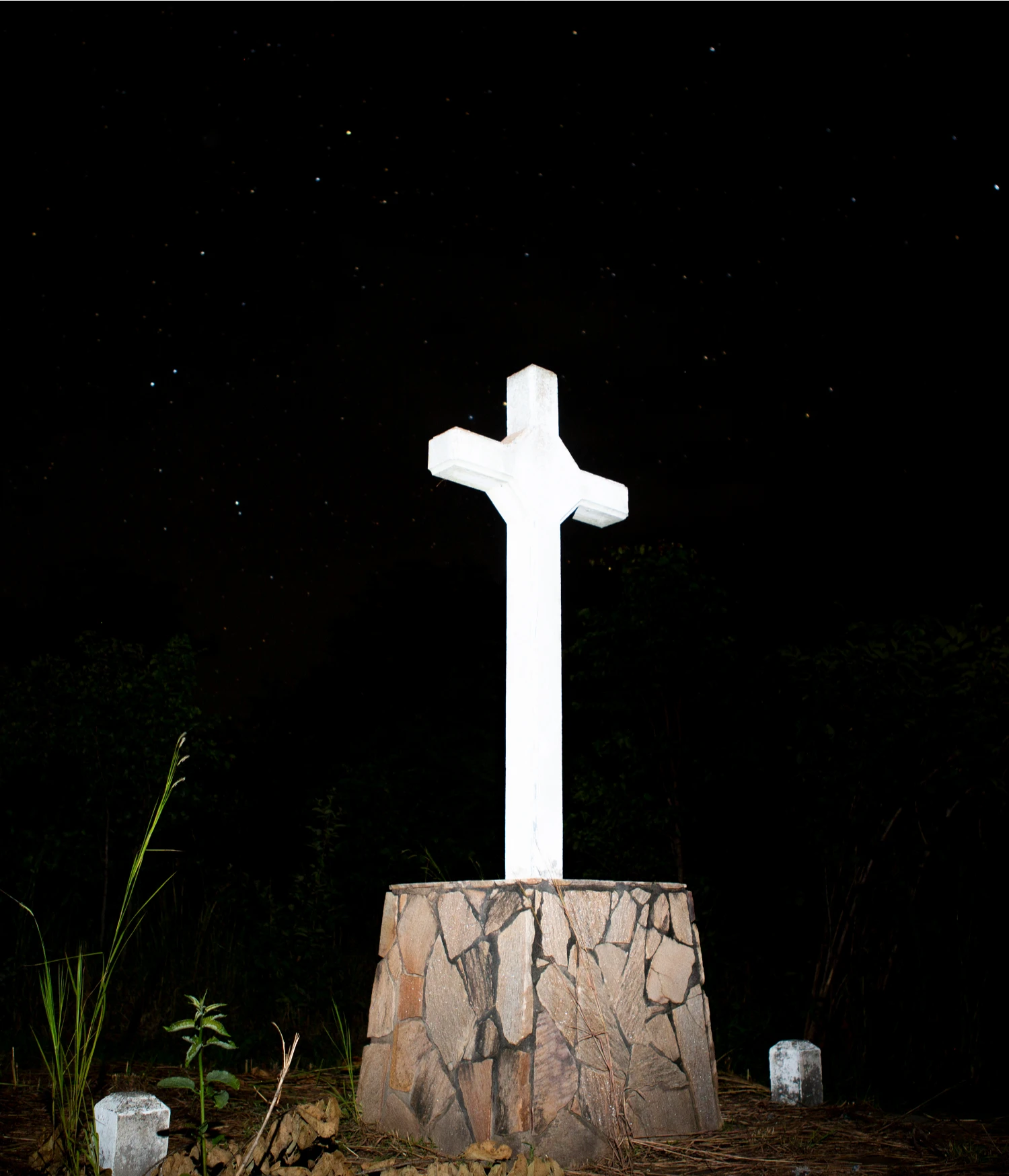 They Still Owe Him a Boat by Jono Terry — A white cross mounted on a stone base rises against a black night sky, its geometric form starkly illuminated.
