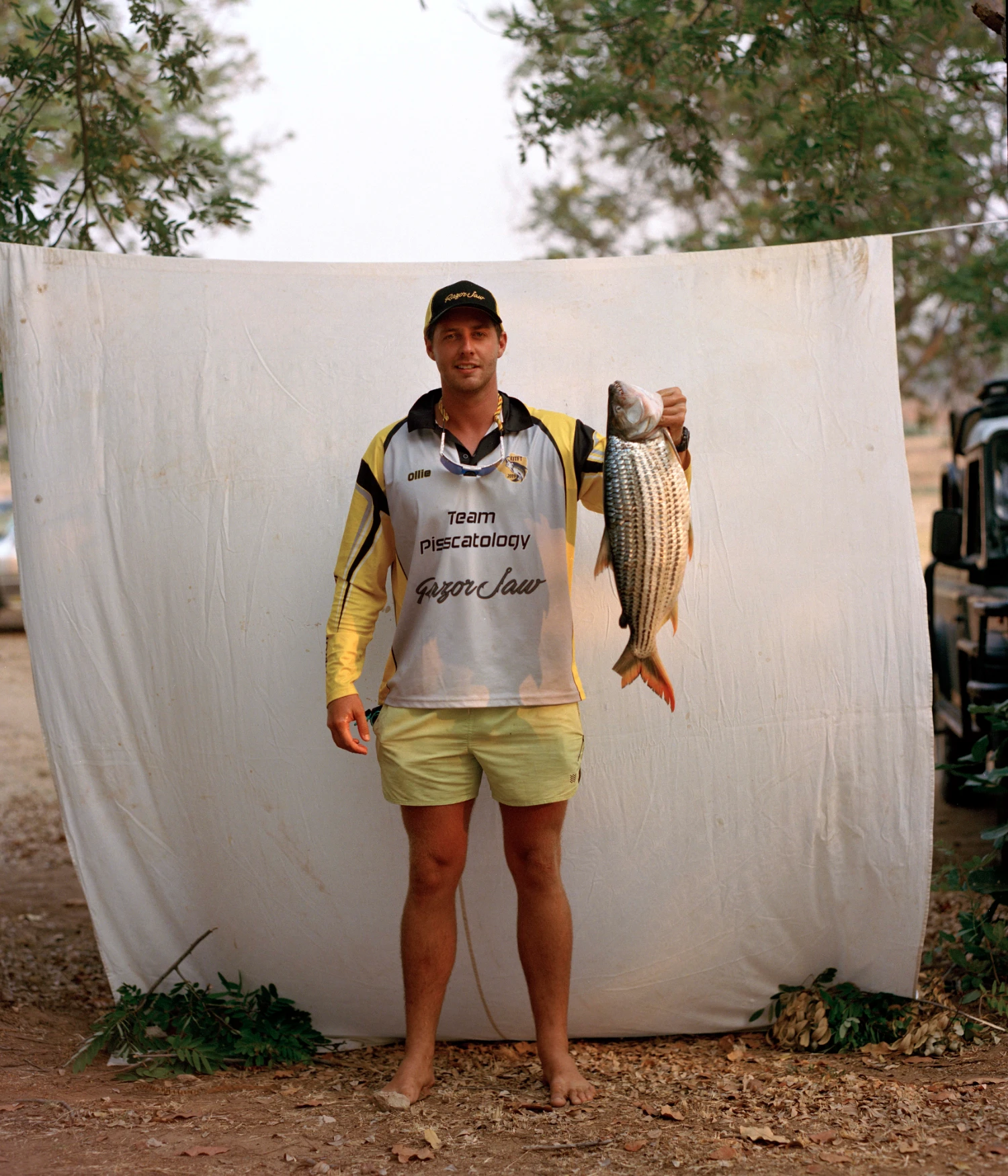 They Still Owe Him a Boat by Jono Terry — A fisherman stands against a white canvas backdrop suspended between trees, holding a freshly caught fish at arm's length.