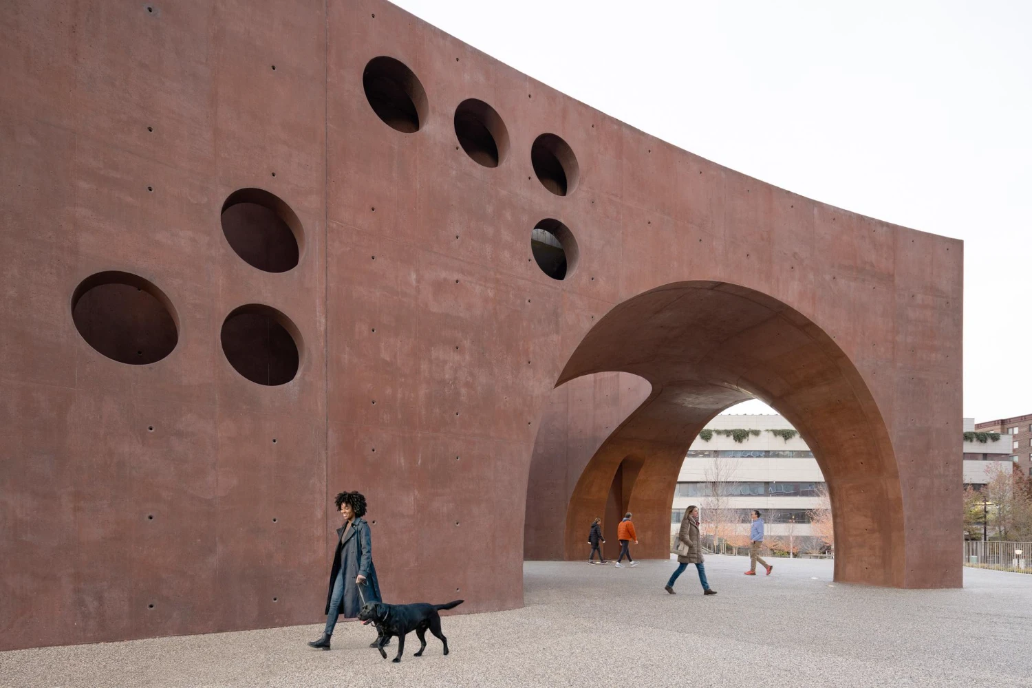 Wagner Park Pavilion by Thomas Phifer — A woman and her black dog walk beneath the Wagner Park Pavilion's monumental twin concrete arches, which frame a view through to the background structure.