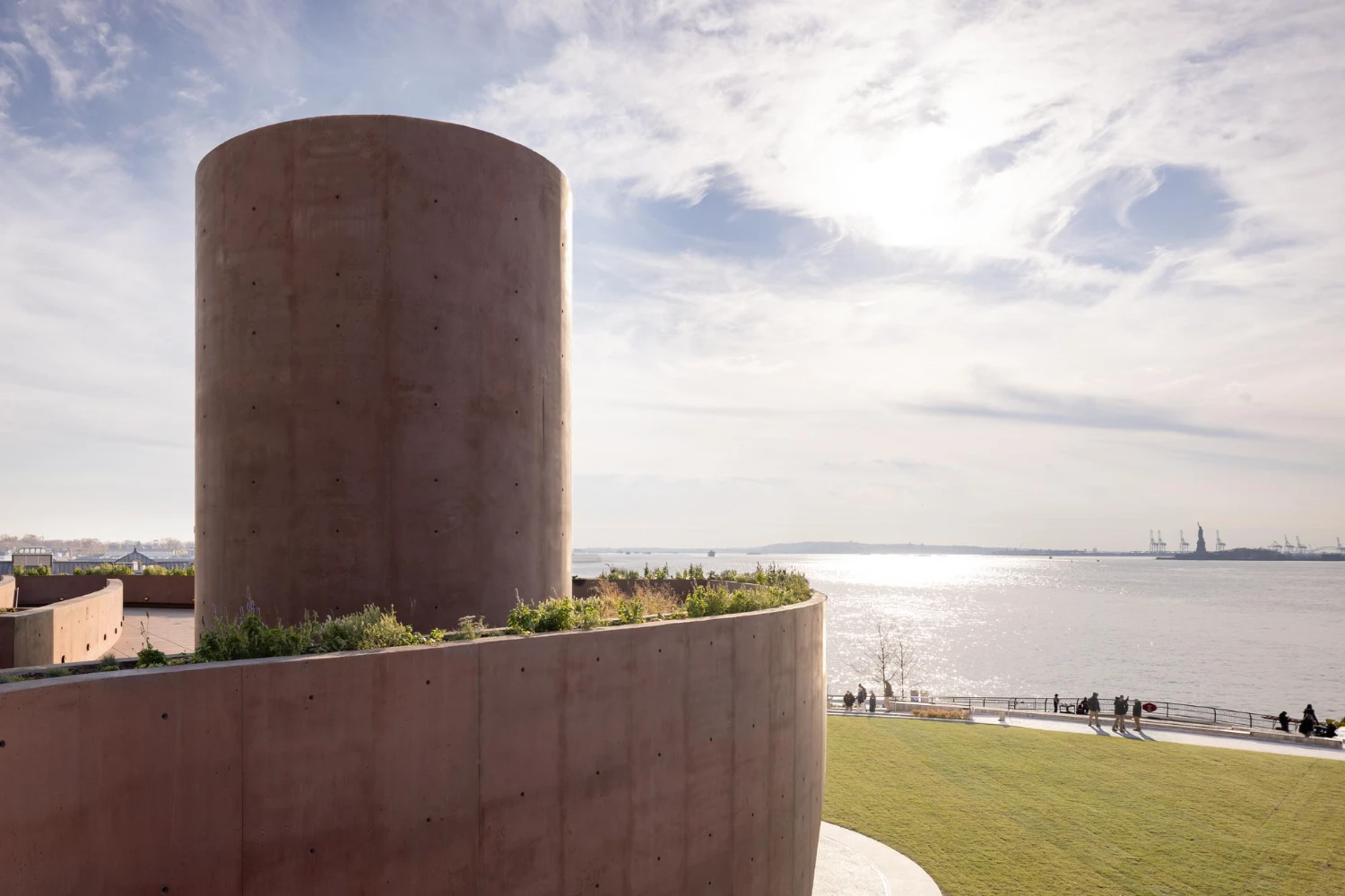Wagner Park Pavilion by Thomas Phifer — A monumental reddish-brown concrete cylinder rises from a sloped lawn, its formwork fastener holes visible across the curved surface.