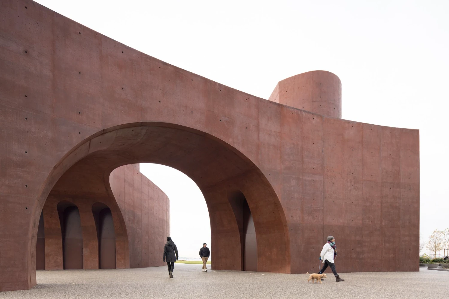 Wagner Park Pavilion by Thomas Phifer — A monumental concrete pavilion dominates the frame with twin curved archways creating a void gateway, their inner surfaces casting deep shadows that...