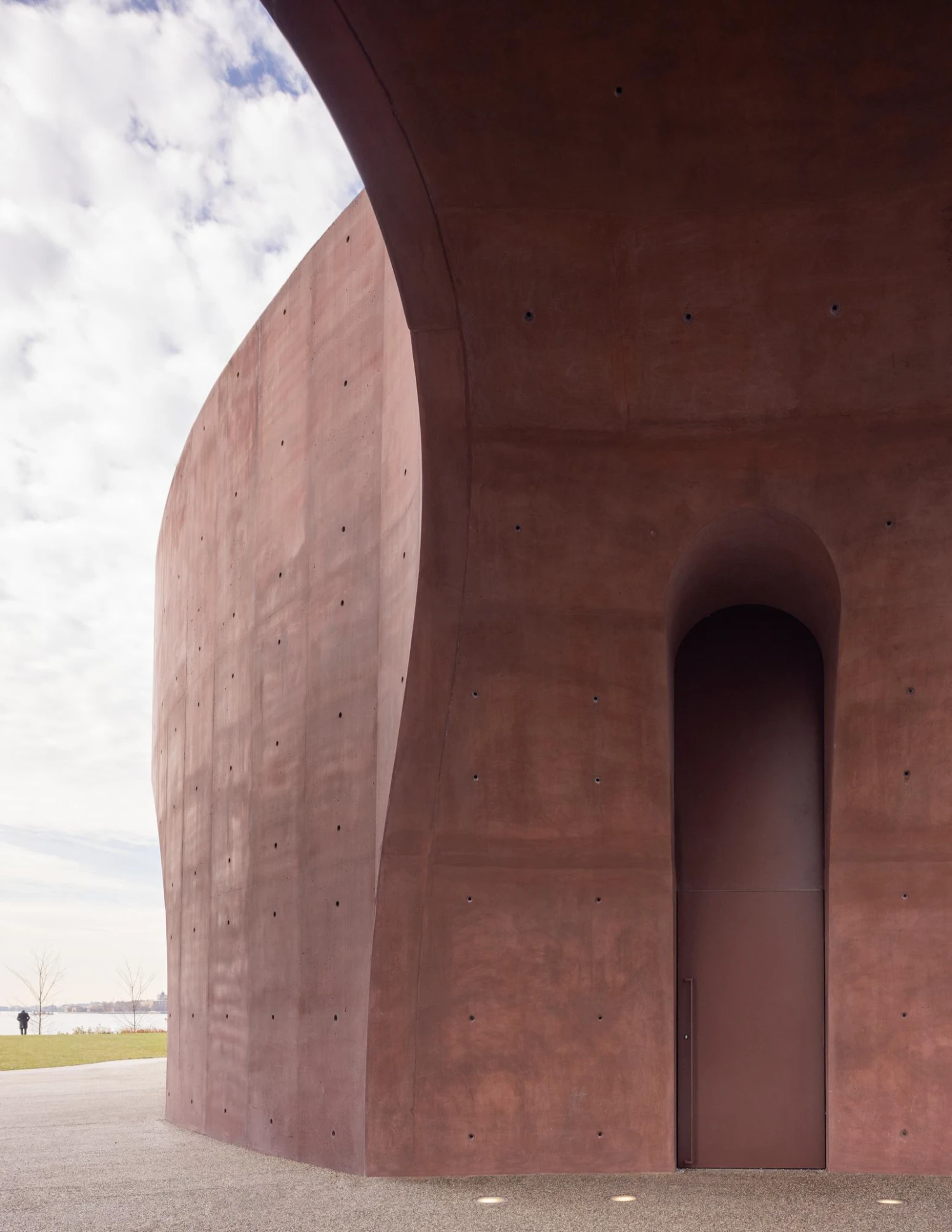 Wagner Park Pavilion by Thomas Phifer — The Wagner Park Pavilion presents a monumental curved concrete facade in reddish-brown, its surface marked by regular fastener holes from formwork.