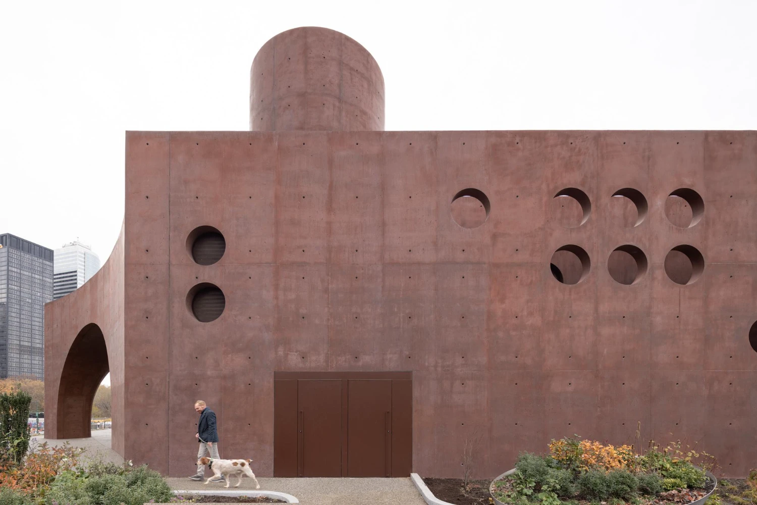 Wagner Park Pavilion by Thomas Phifer — A monumental reddish-brown concrete pavilion dominates the frame, its mass punctuated by geometric voids that structure the composition.