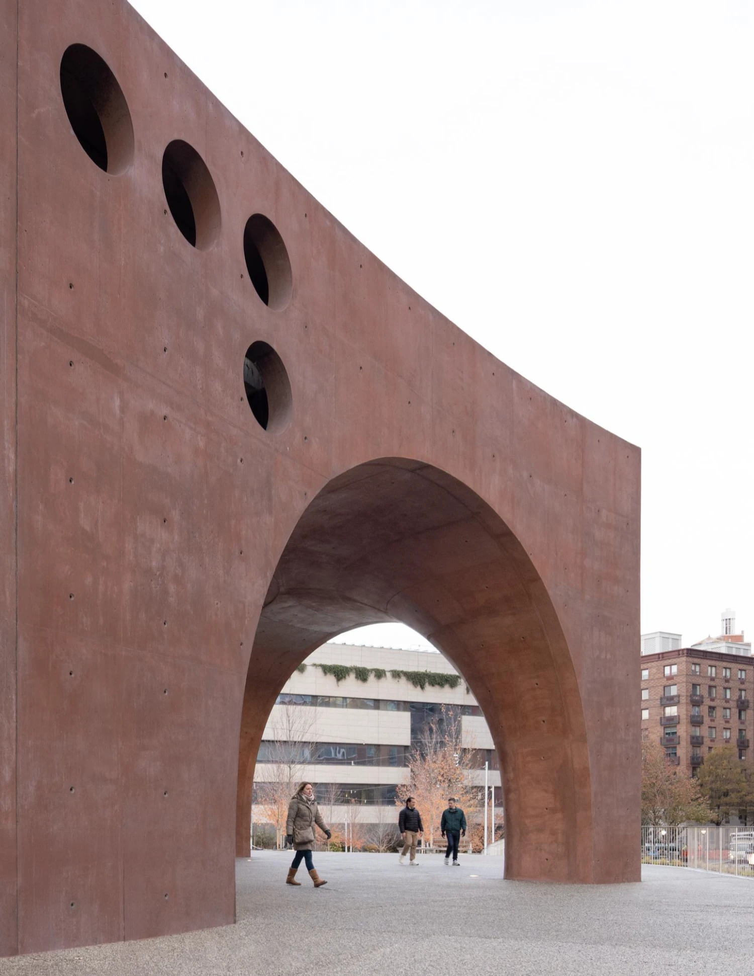 Wagner Park Pavilion by Thomas Phifer — A monumental curved concrete archway dominates the composition, its reddish-brown surface marked by visible formwork fastener holes across the upper wall plane.