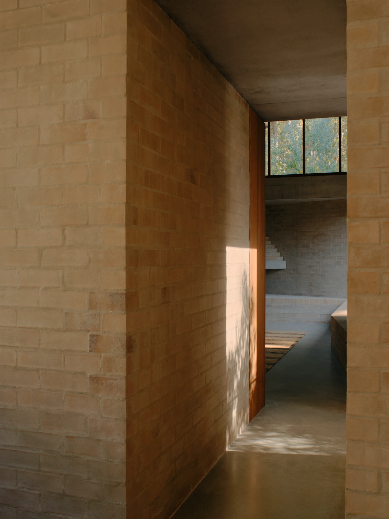 Stuartholme House by Nielsen Jenkins + Michael Lumby — A timber-framed doorway opens onto a sunken interior passage, where brick walls rise in stepped planes toward a concrete soffit ceiling.