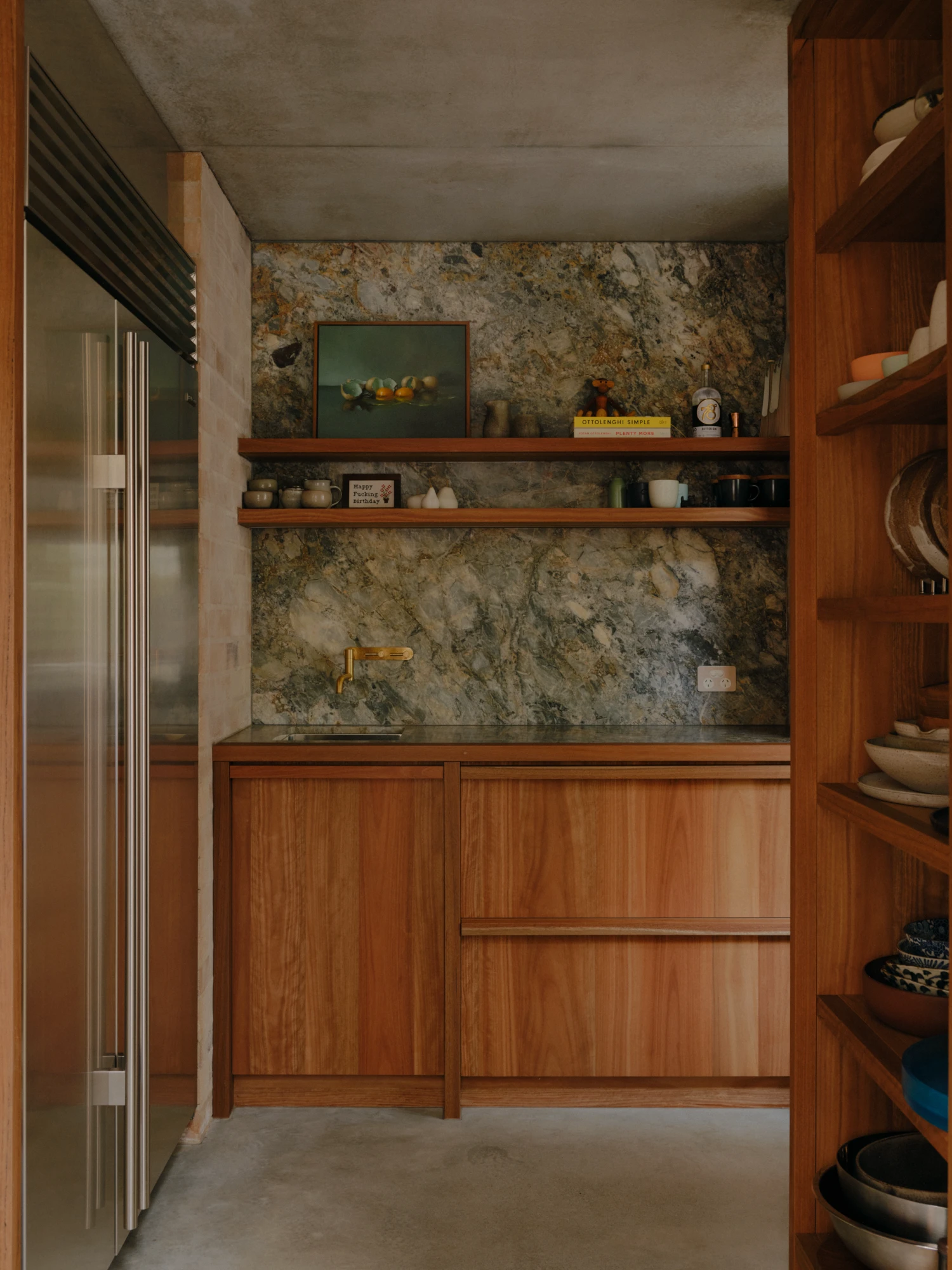 Stuartholme House by Nielsen Jenkins + Michael Lumby — A compact wet kitchen anchors the gully-side residence, its warm timber cabinetry contrasting sharply with the mottled granite-effect stone wall rising behind.