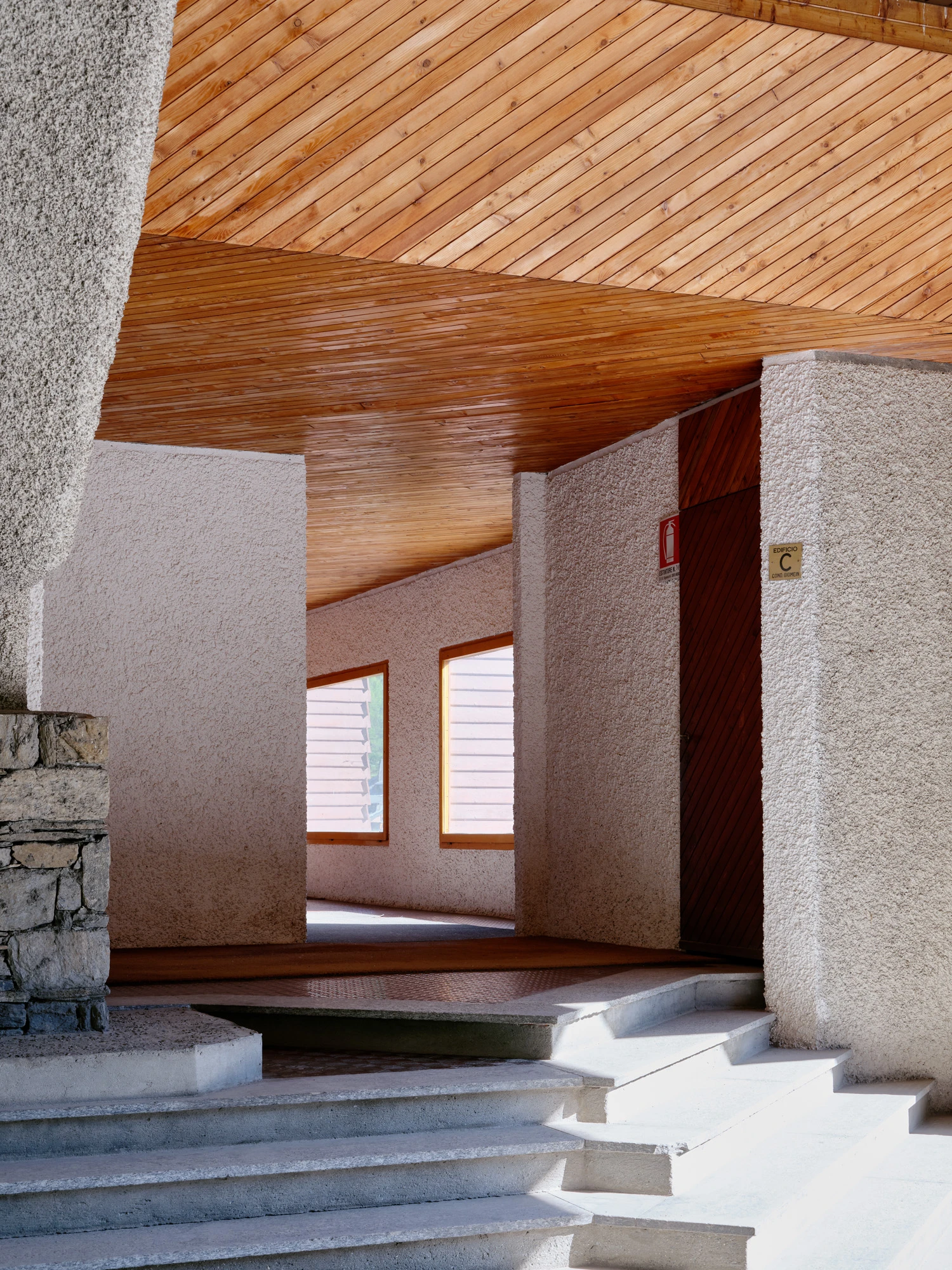 An Interior at Giomein by co.arch — A passage framed by render-finished walls and timber-lined ceiling, where the pitched roof geometry becomes volumetric.