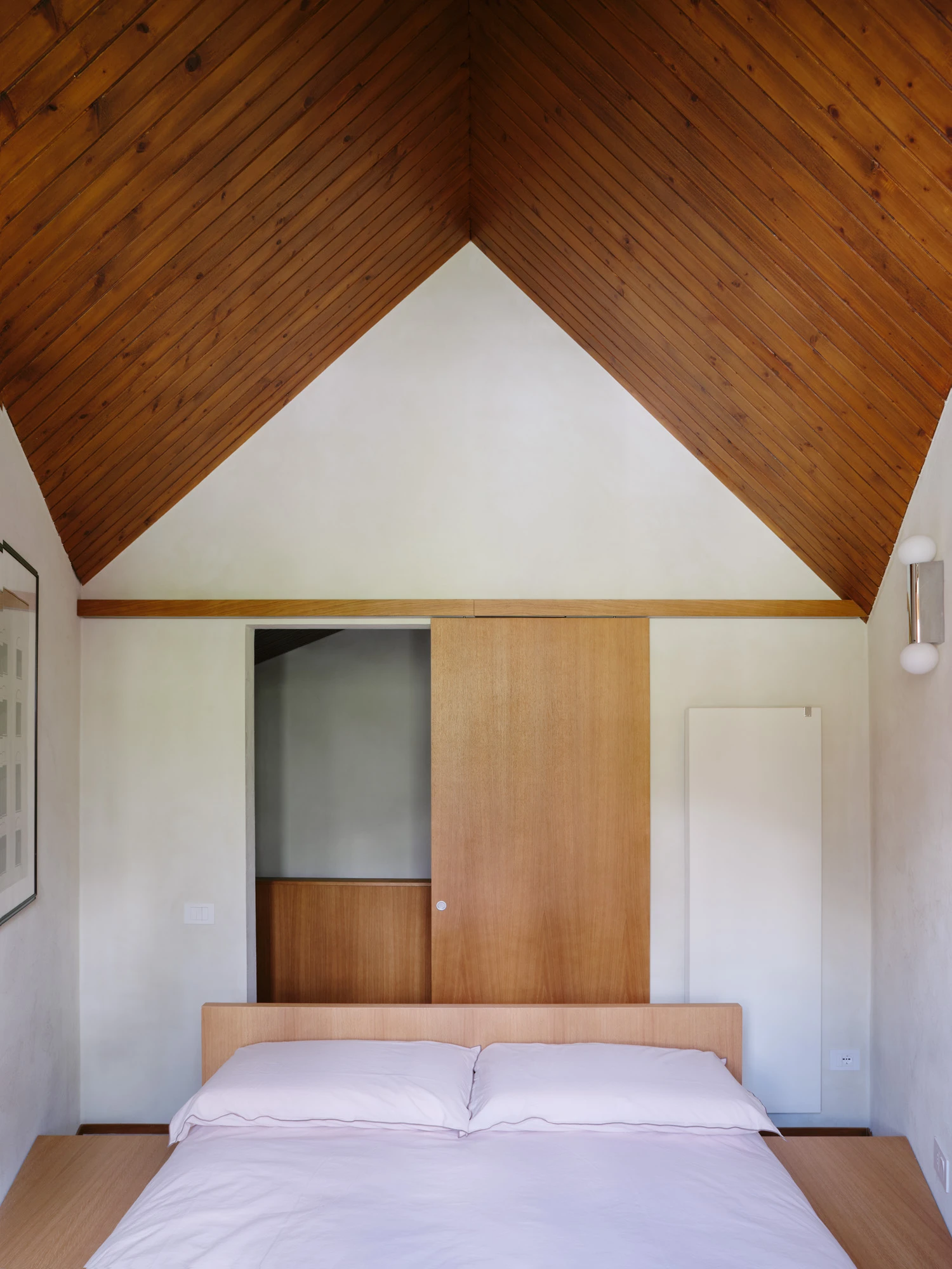 An Interior at Giomein by co.arch — A bedroom interior framed from the foot of the bed, revealing the project's essential gesture: the exposure of the pitched roof's timber structure.
