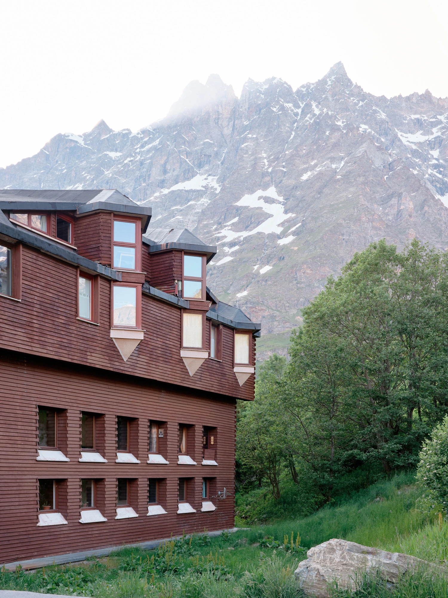 An Interior at Giomein by co.arch — The red-brown timber-clad facade of Giomein rises against snow-capped Alpine peaks, its eccentric geometry evident in the steeply pitched roof planes and...